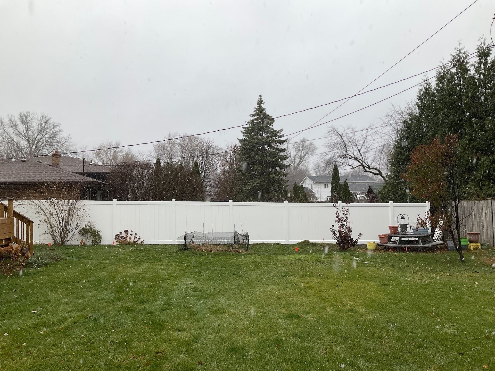 Snow falling in a backyard with a white fence, green grass, and trees under a cloudy sky.