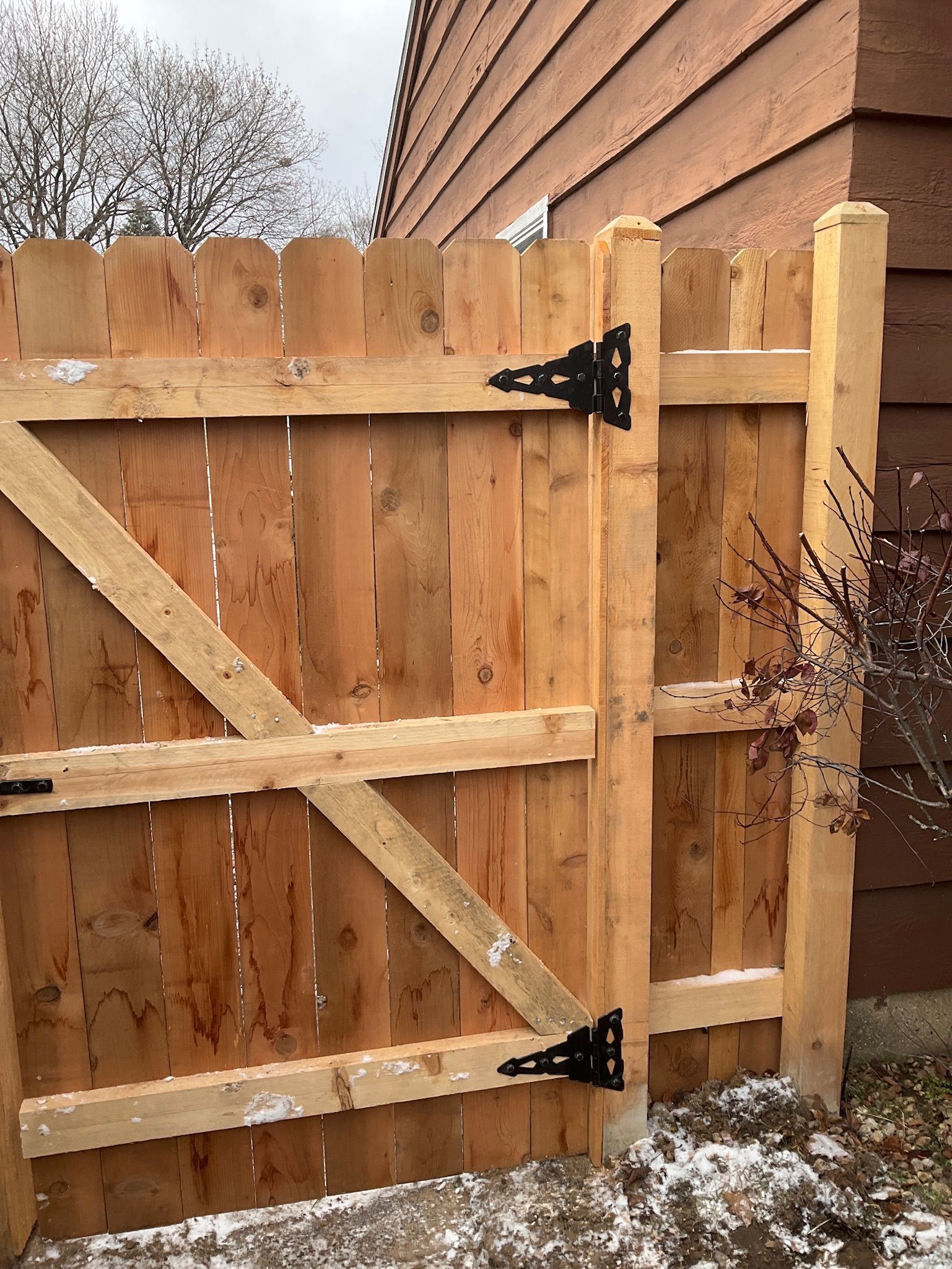 Wooden gate with black hinges, next to a brown siding wall, and a bit of snow.