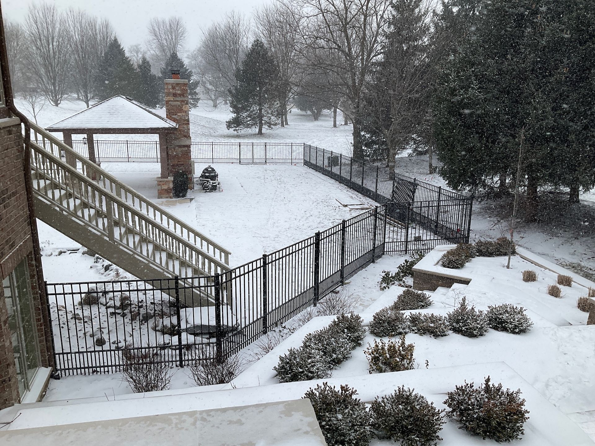 Snowy backyard scene with a deck, gazebo, and black fence. Trees and bushes covered in snow.