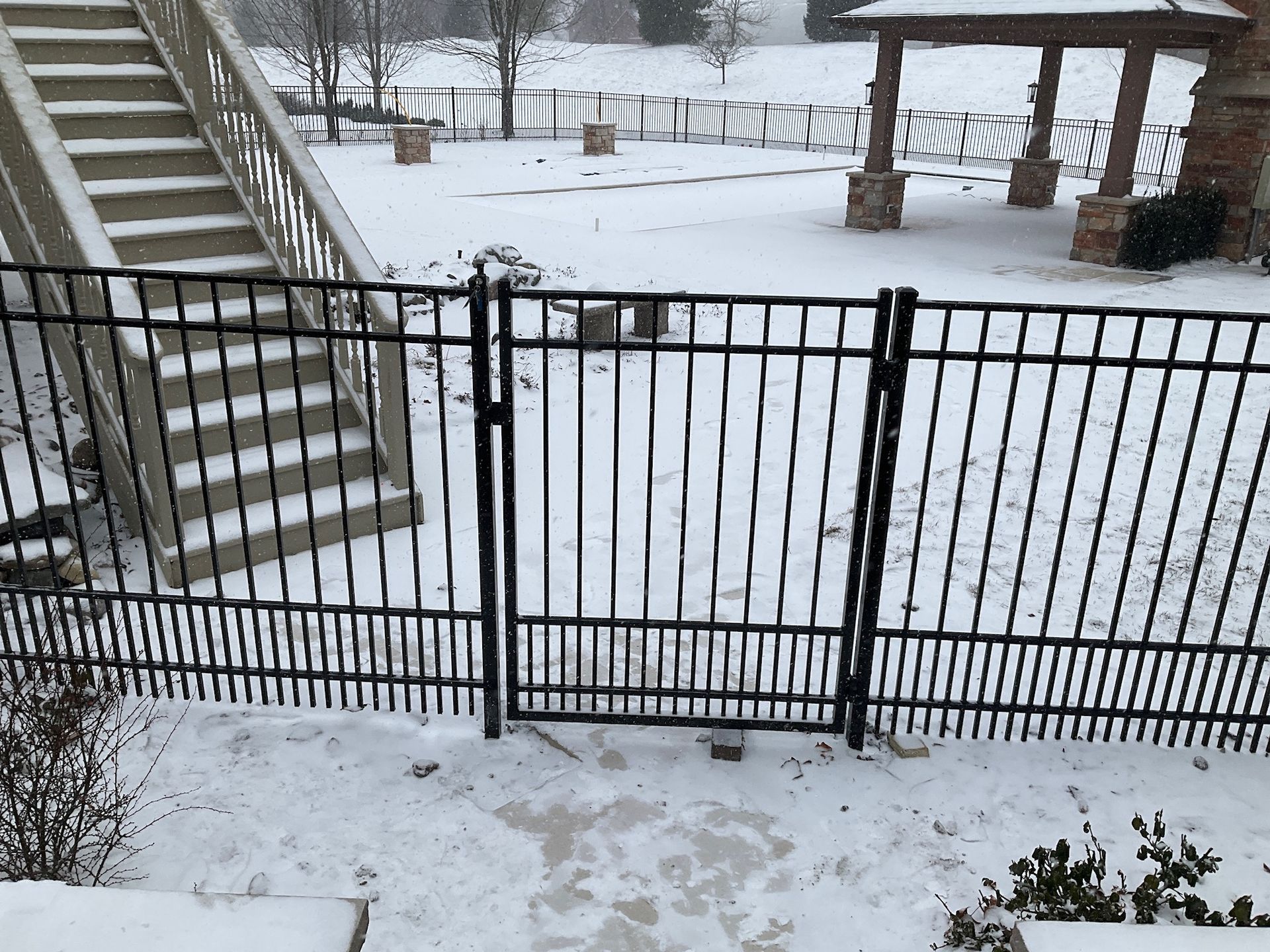 Snowy backyard scene with black metal fence and gate. Stairs and a gazebo in the background.