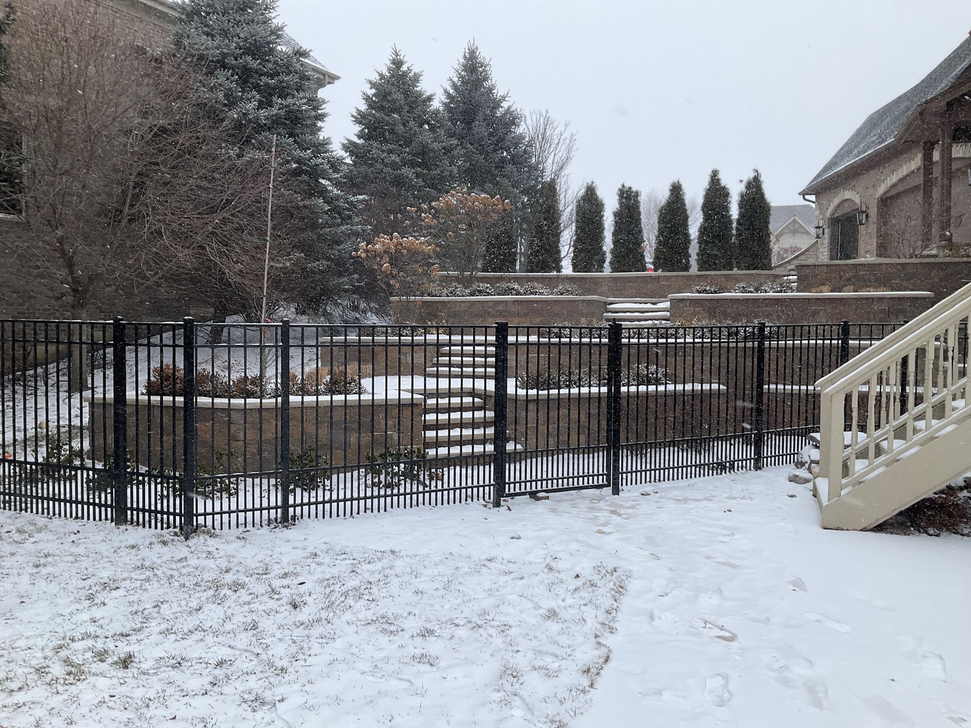 Snowy backyard scene with black fence, stone walls, and evergreen trees.