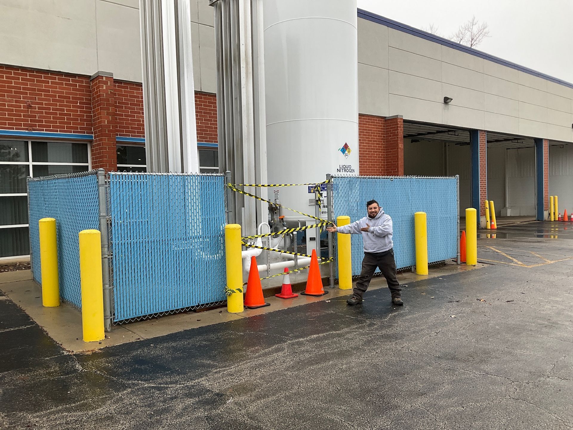 Man standing near a large industrial tank, gesturing. Blue fence, caution tape, and safety cones surround the tank.