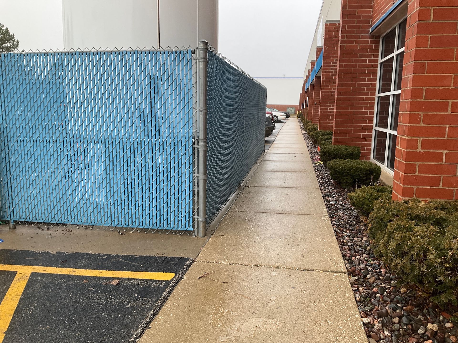 Blue chain-link fence beside a concrete walkway, brick building on right, gray sky.