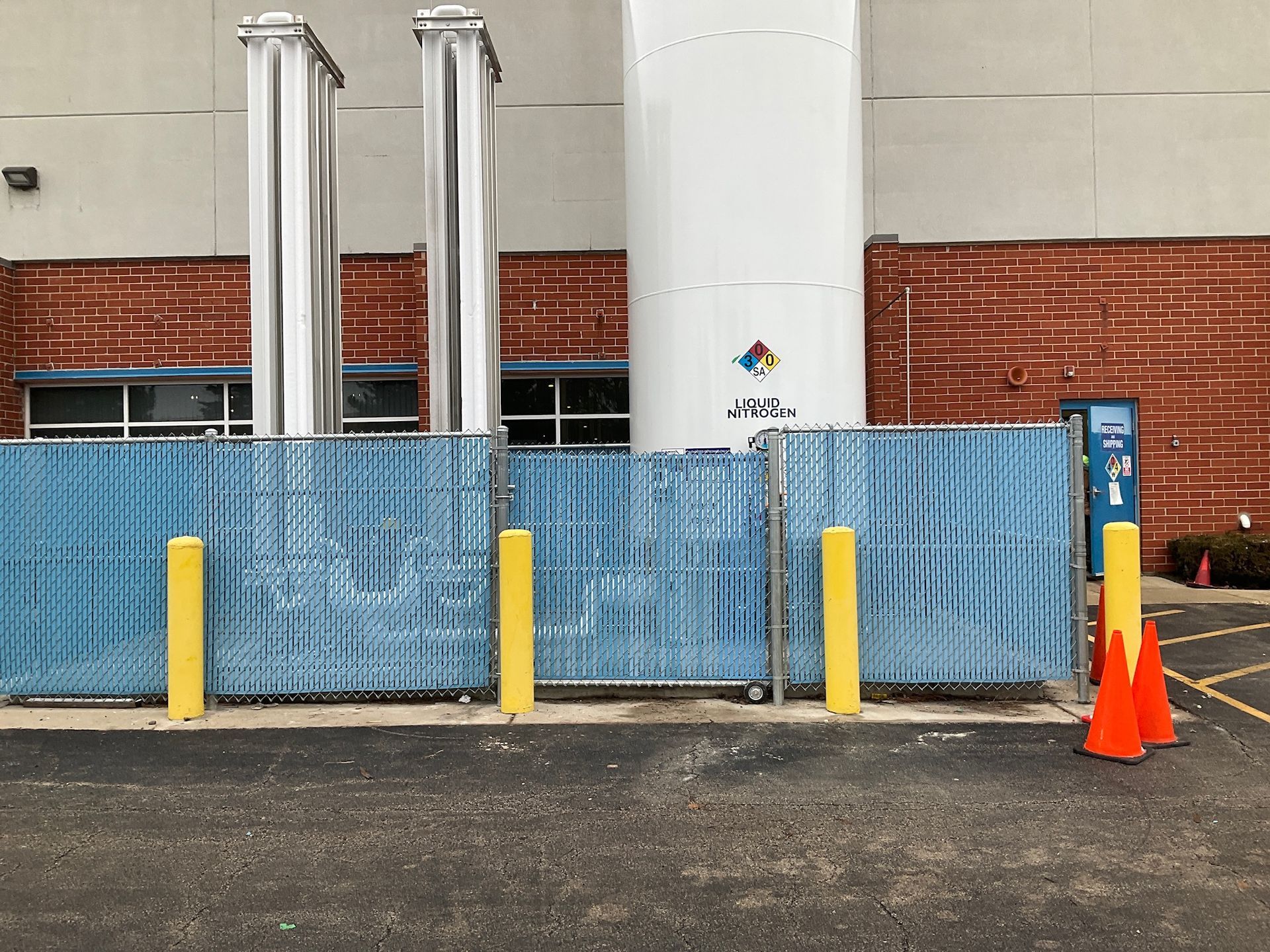 Blue fence in front of industrial structures. Yellow posts and orange cones in parking lot.