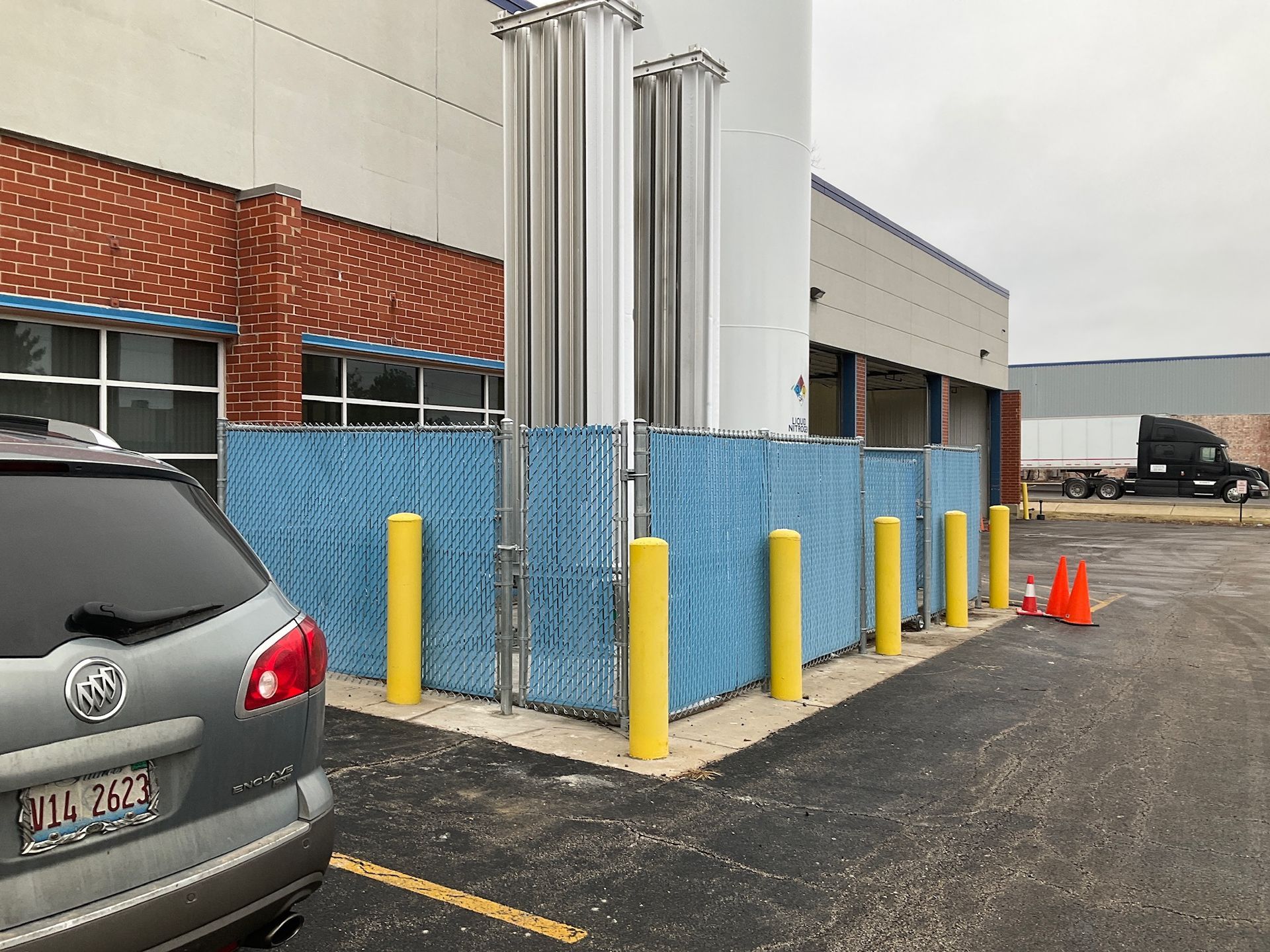 A silver car parked near a building with tall silver tanks behind a blue chain-link fence.