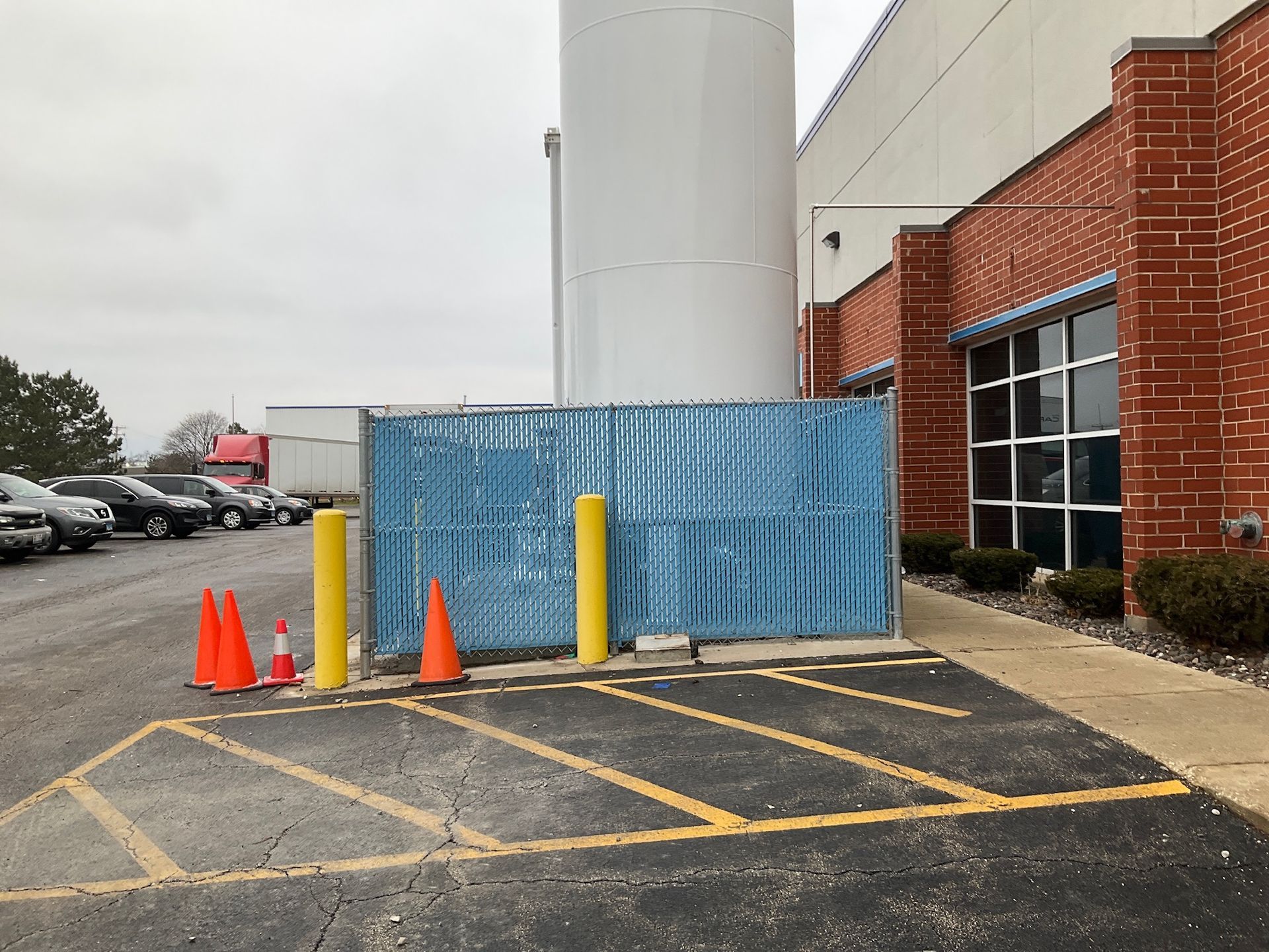 A blue chain-link fence blocks an area next to a white silo and brick building; traffic cones and yellow posts in the foreground.