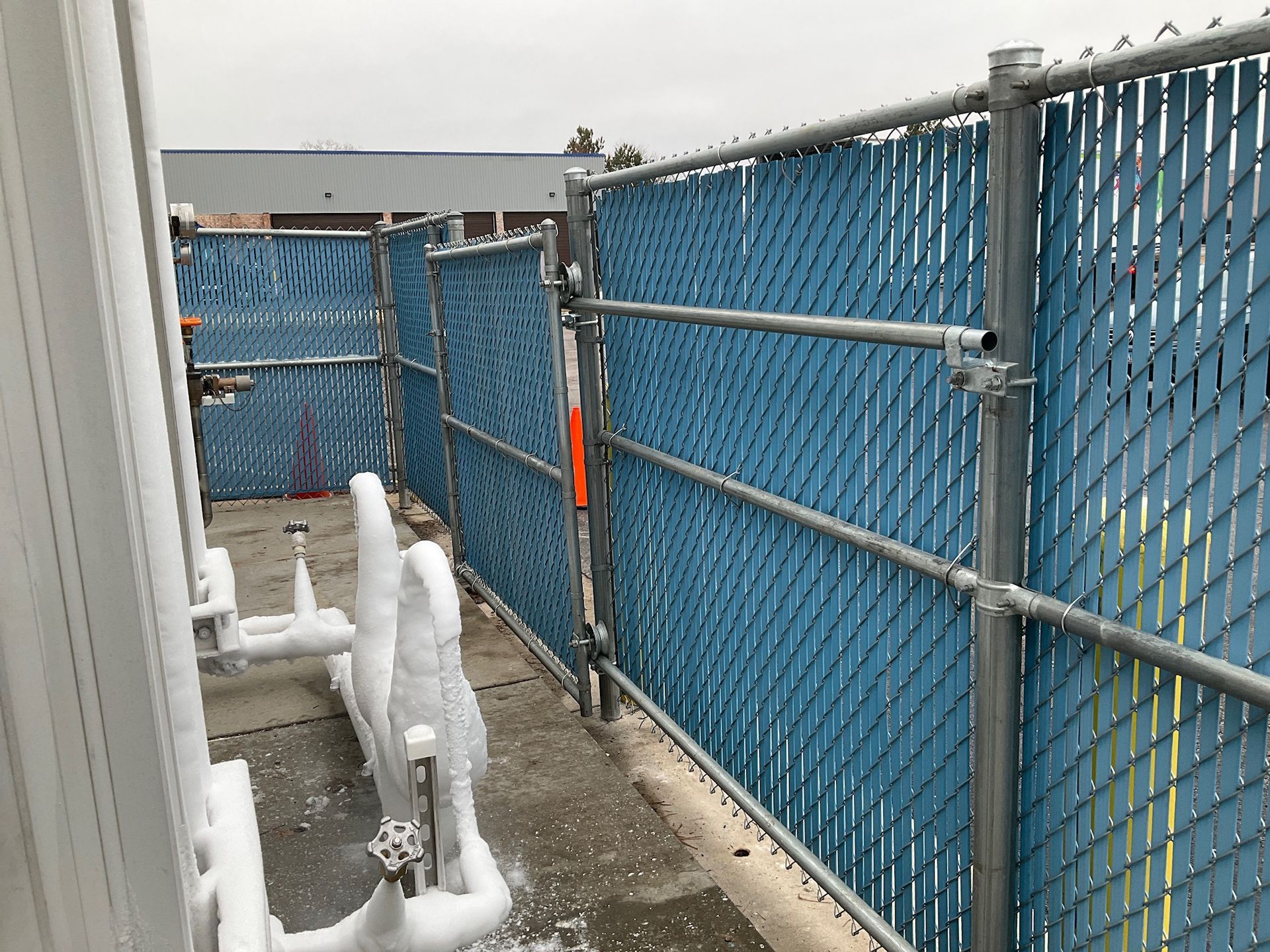 Blue chain-link fence with privacy slats, snow-covered metal pipes, and a gate in an outdoor area.