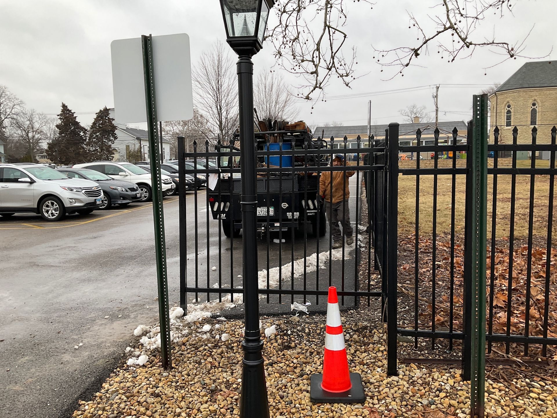 Black truck at black metal fence gate; orange cone, street lamp, parked cars, snow.