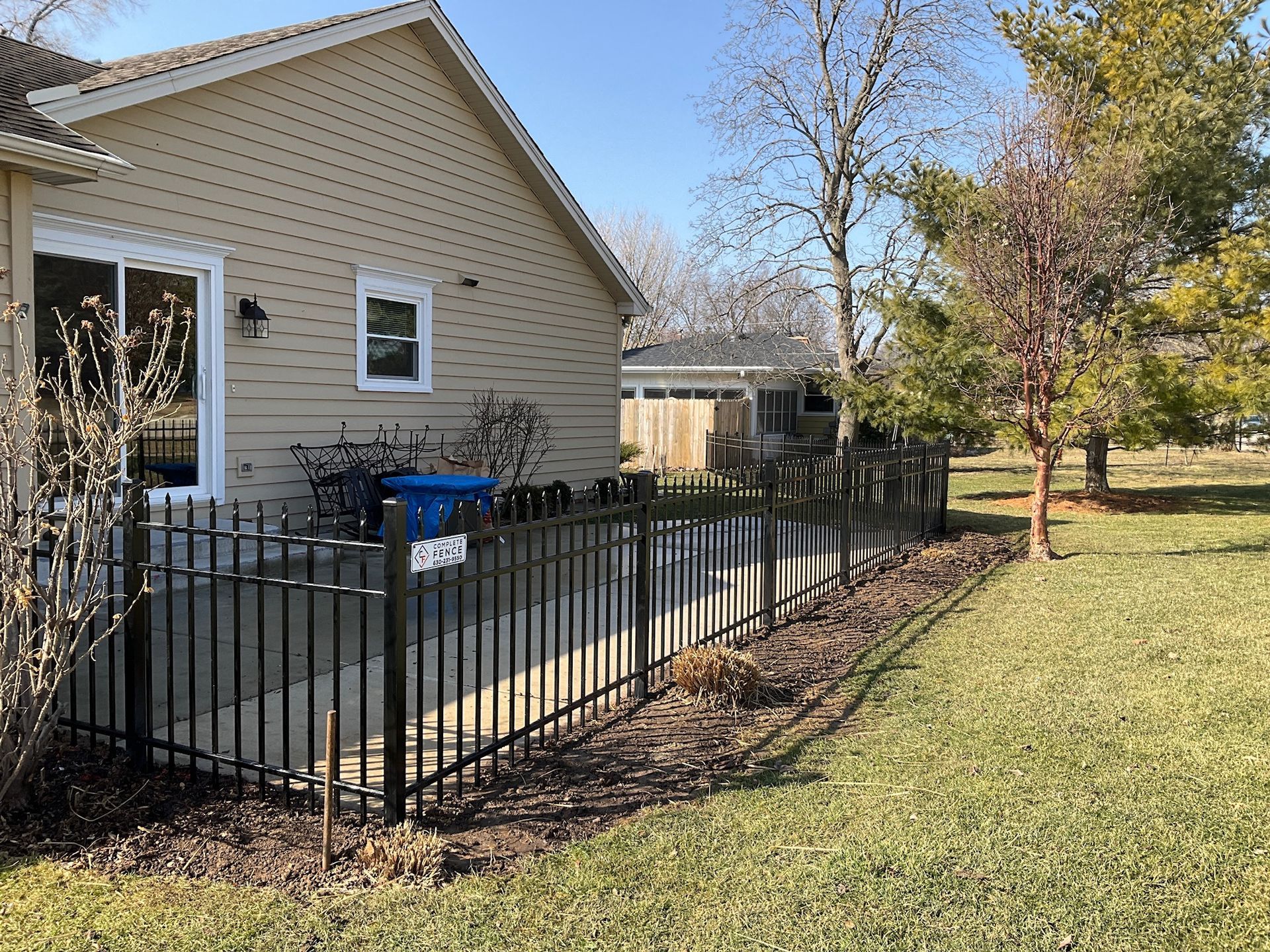 A black metal fence surrounds a concrete patio next to a beige house, sunny day.