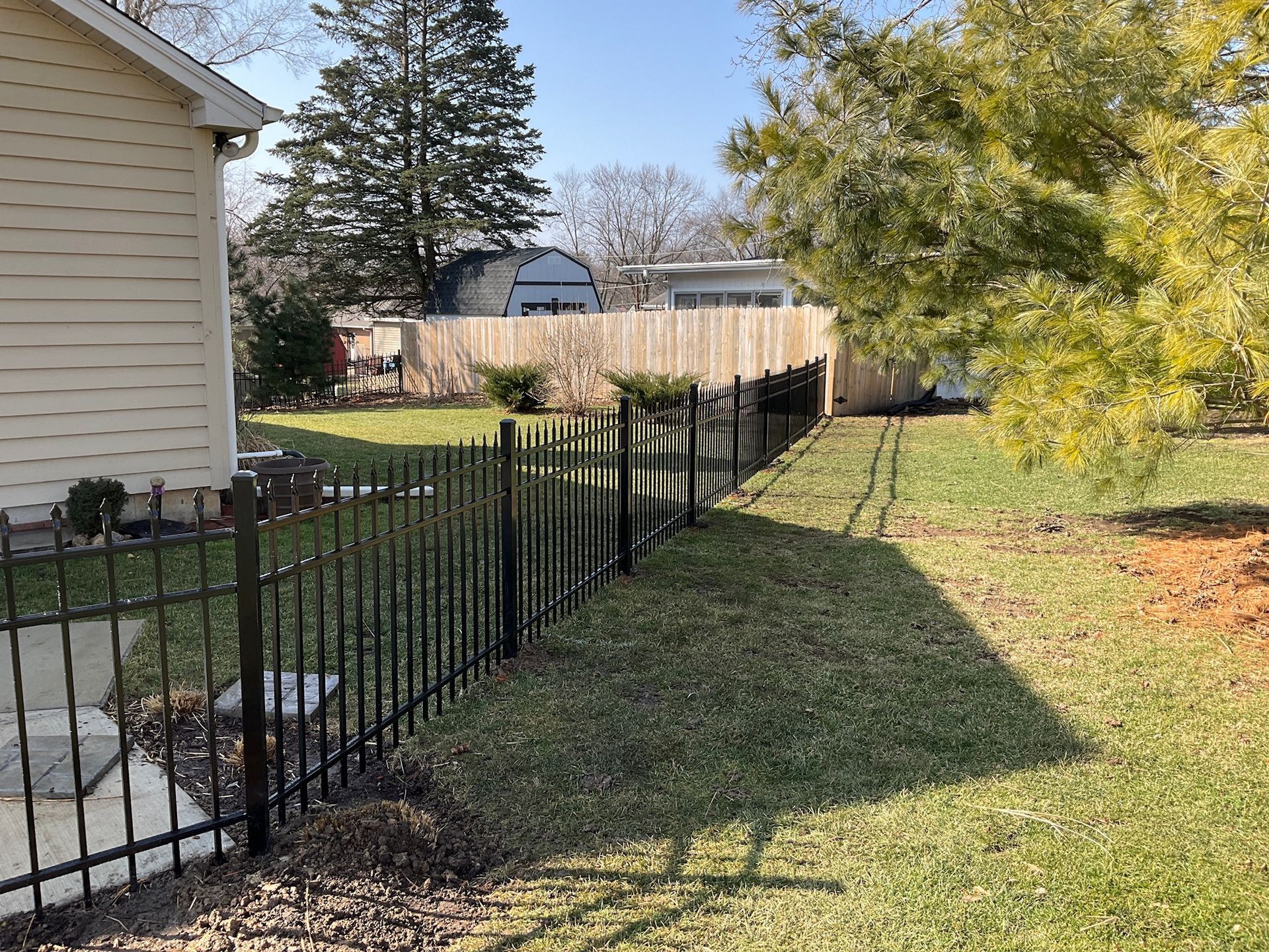 Black metal fence along a grassy yard, beside a house with a wooden fence in the background. Sunny day.