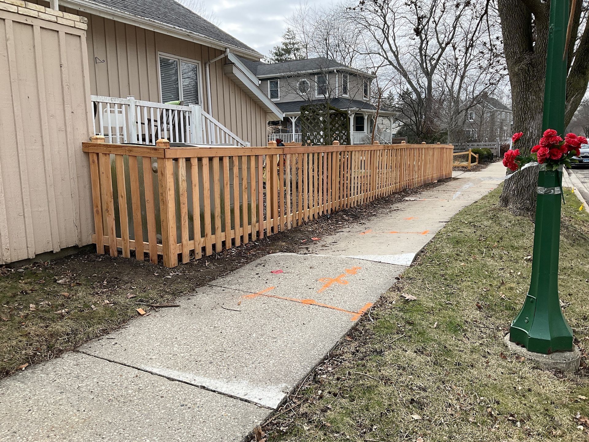 Wooden fence alongside a sidewalk, next to a building and tree.