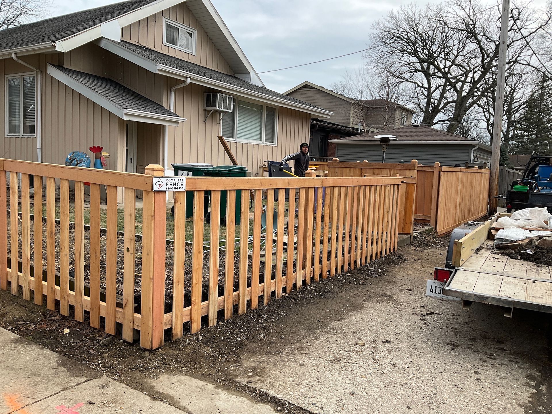 Wooden fence being built in front of a tan house. Workers and a trailer are nearby.