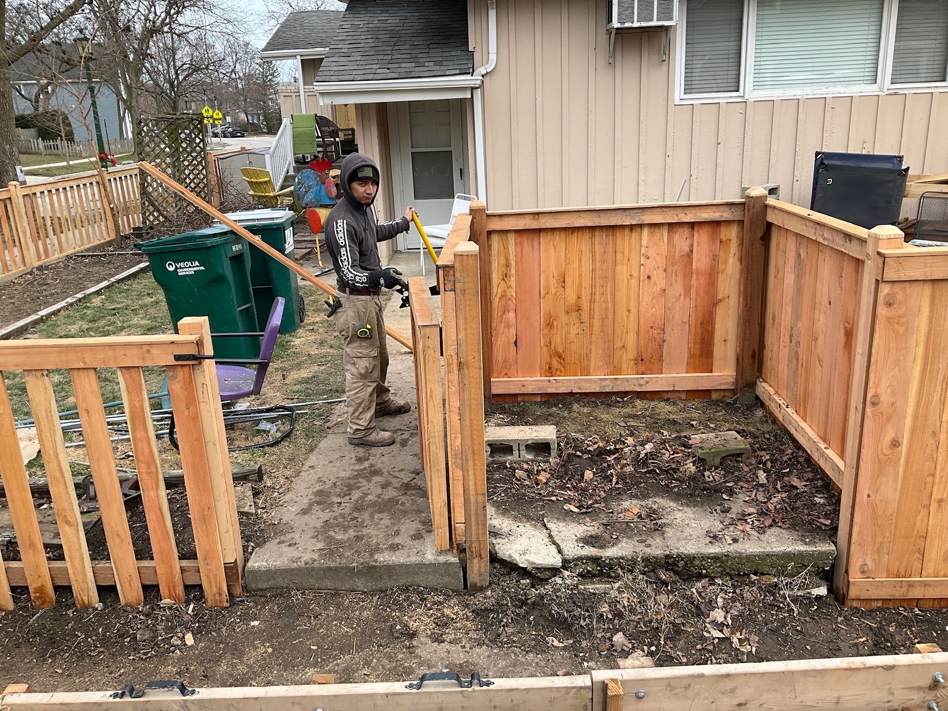 Man measures fence during construction, outdoors near a house with a green trash bin.