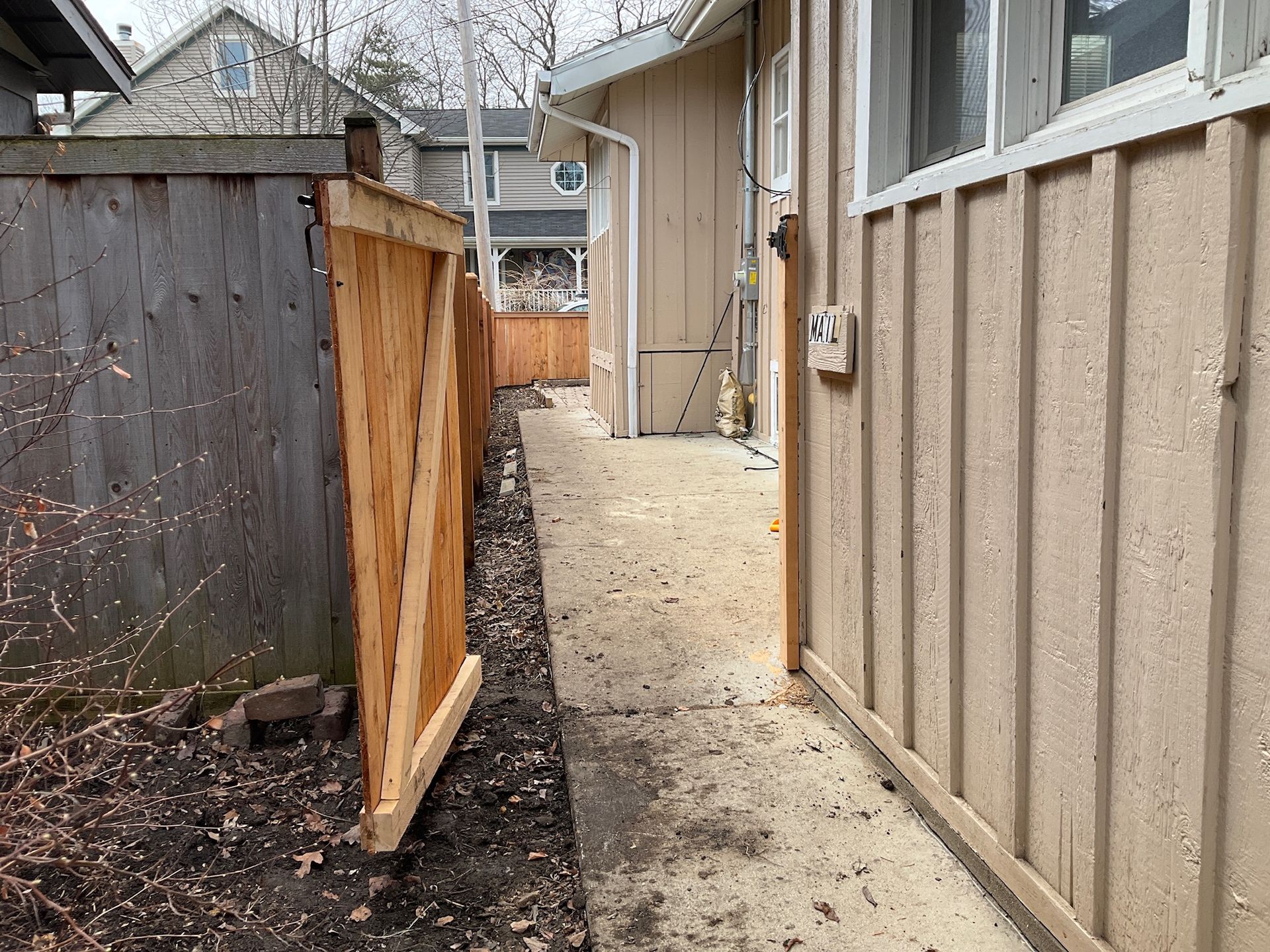 Narrow concrete walkway between a wooden fence and a beige house wall.