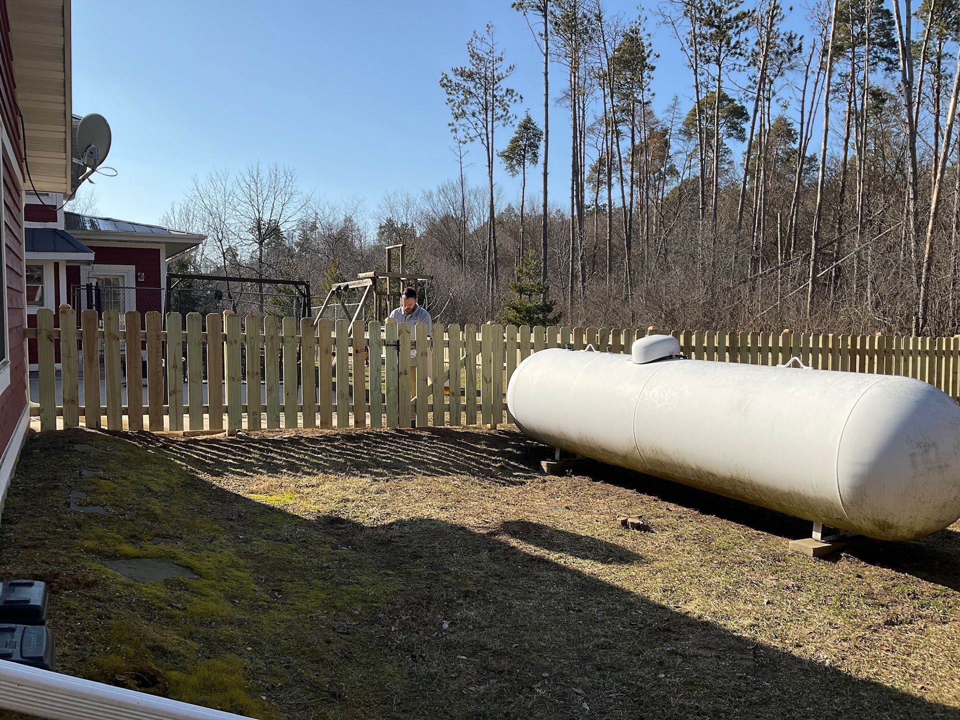 A wooden picket fence surrounds a yard with a large white propane tank.  A building is on the left; trees are in the background.