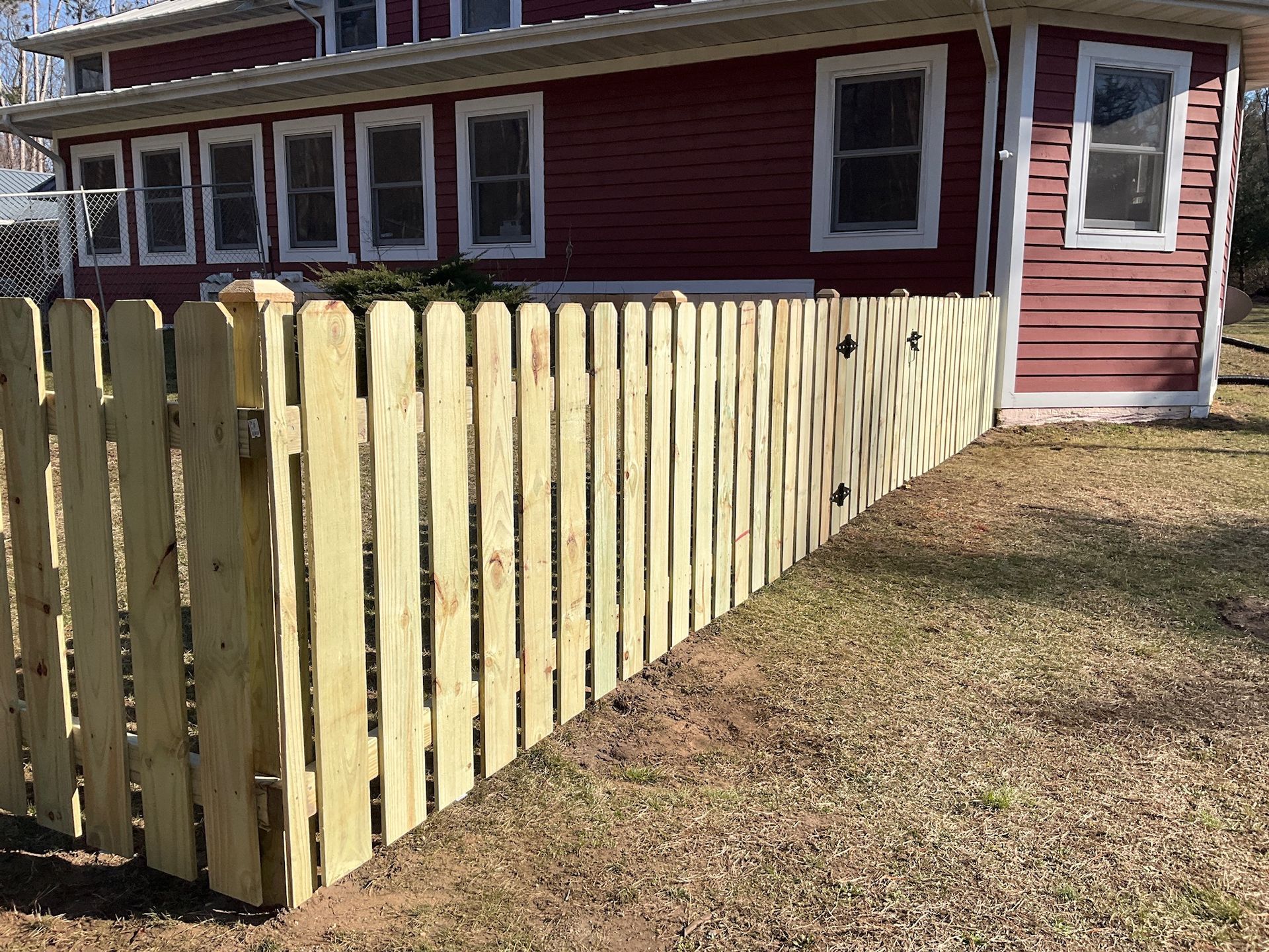A newly constructed, light-yellow picket fence with a gate in front of a red building with white-framed windows.