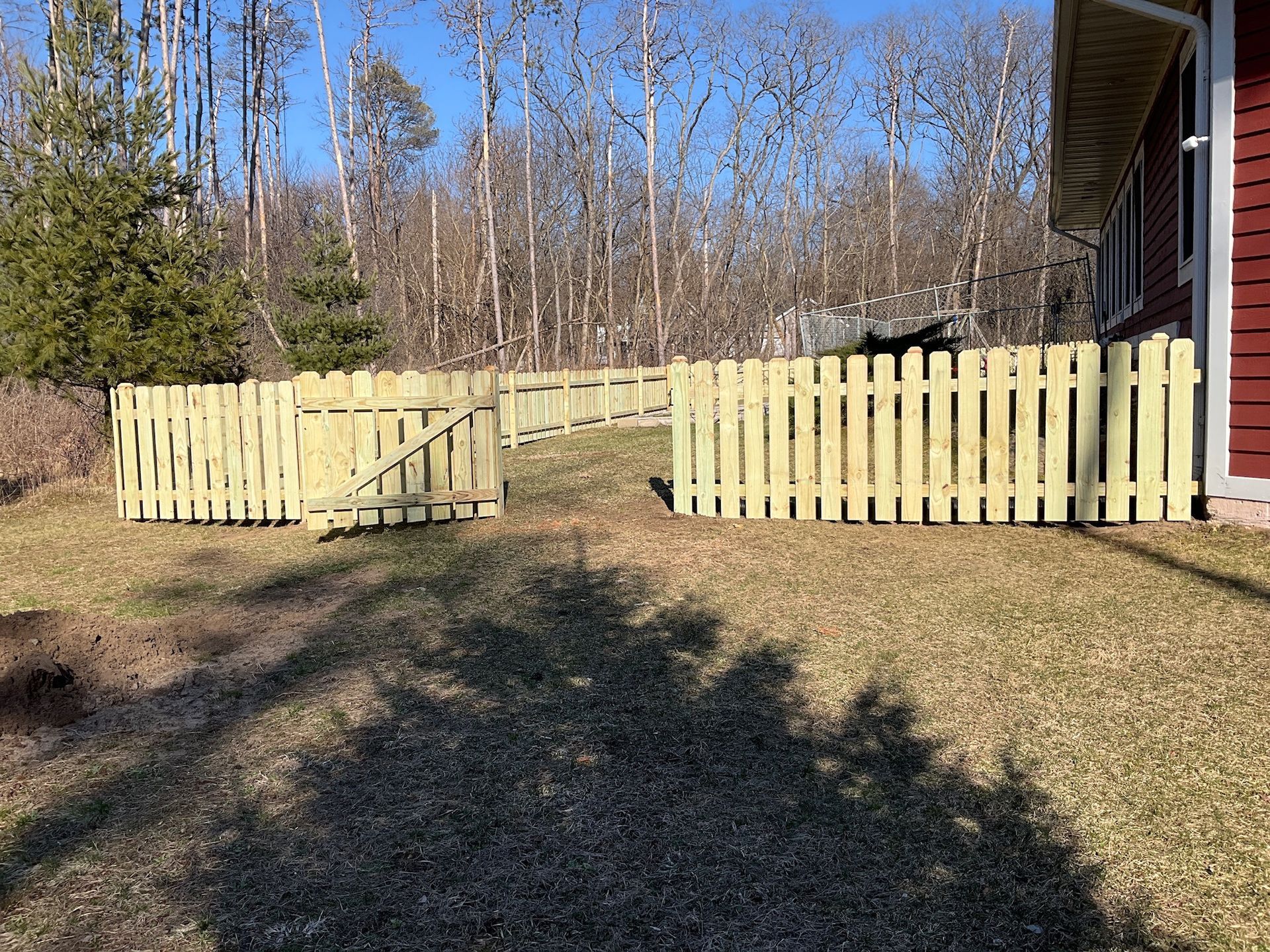 Wooden picket fence with gate in a yard, leading to a wooded area, next to a red house.