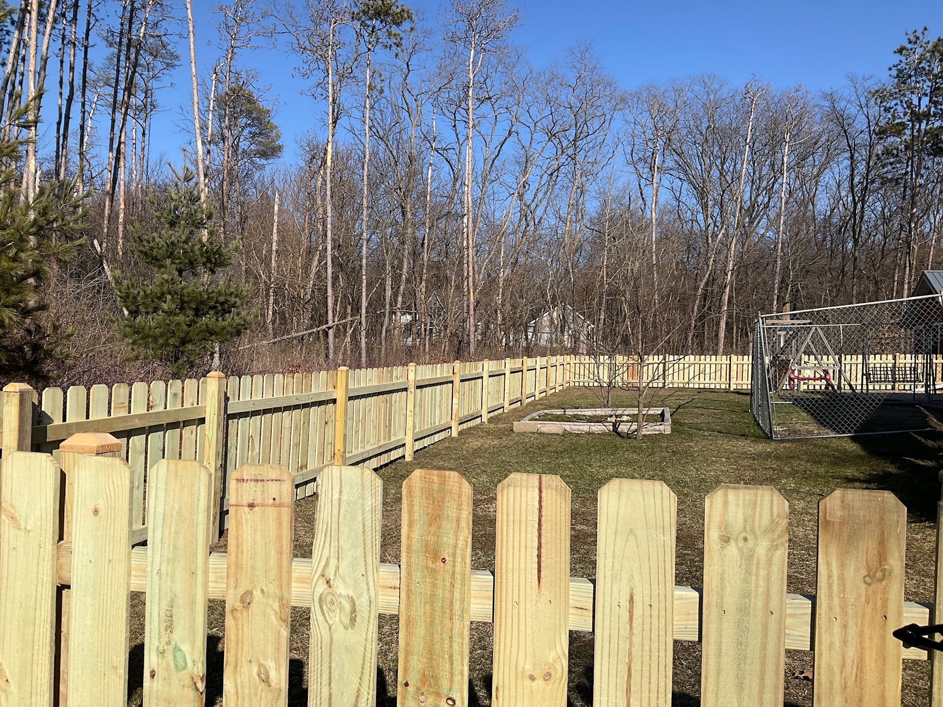 Wooden picket fence in a backyard with trees in the background. Sunny day.