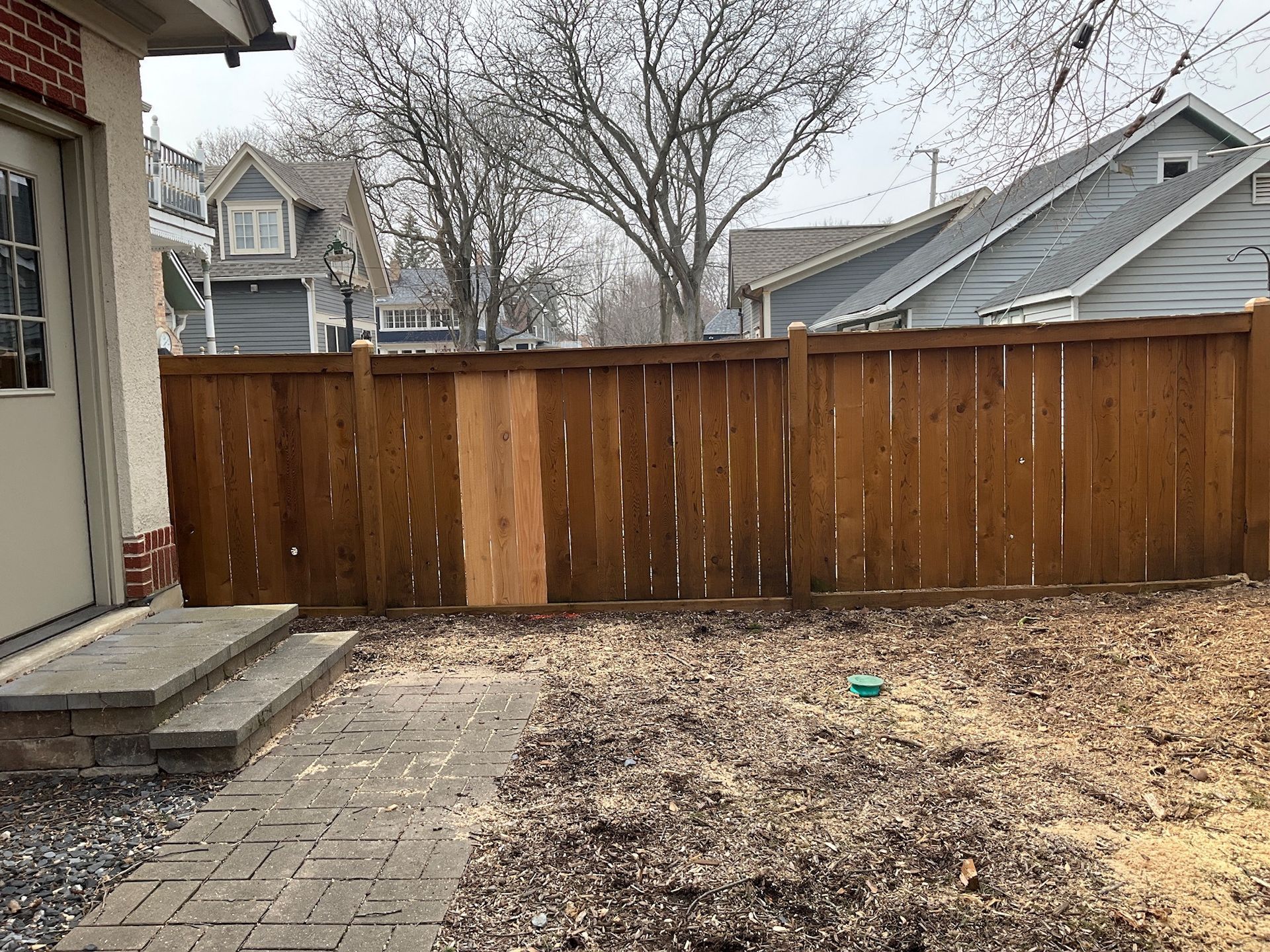 Backyard view of a wooden fence, brick path, and steps leading to a beige door.