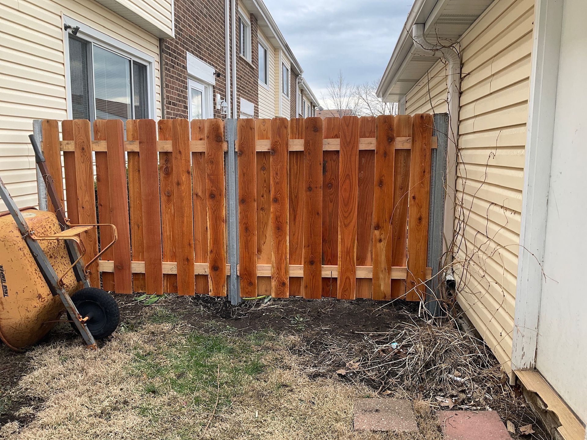 Wooden fence between two buildings; a wheelbarrow sits to the left on the grass.