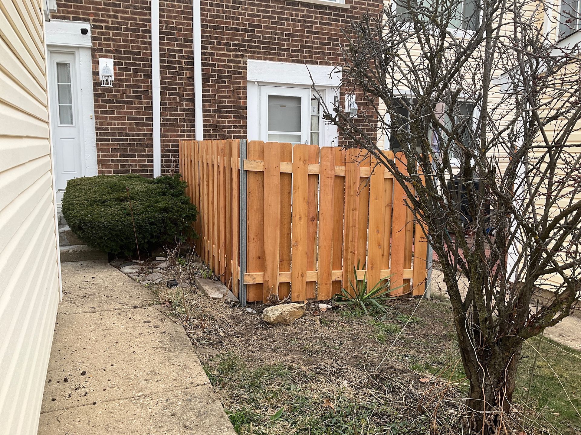 Exterior view of a home with a wooden fence. Pathway leads to a door, bush, and small tree.