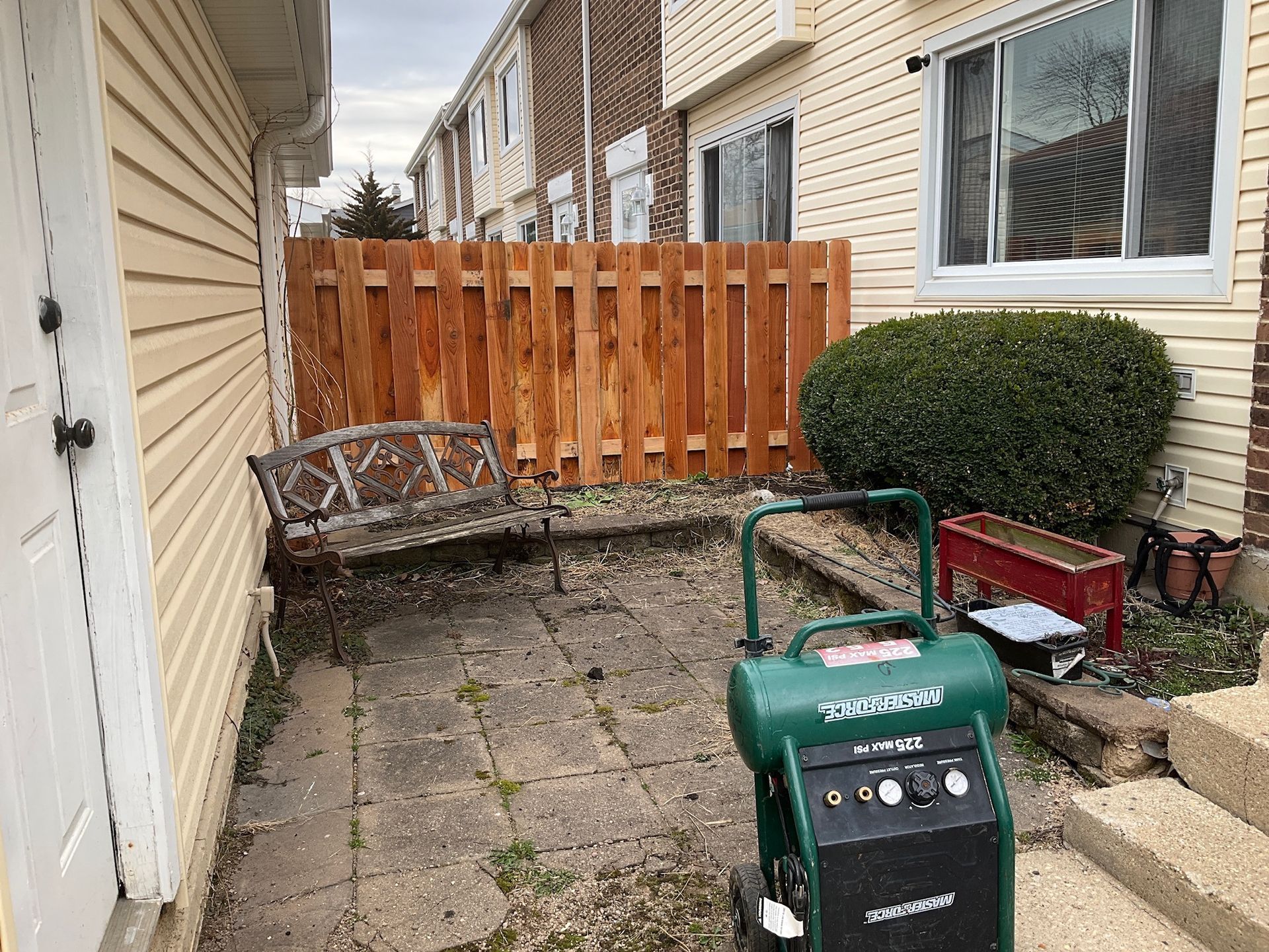 A small backyard patio with a wooden fence, an old bench, and an air compressor.