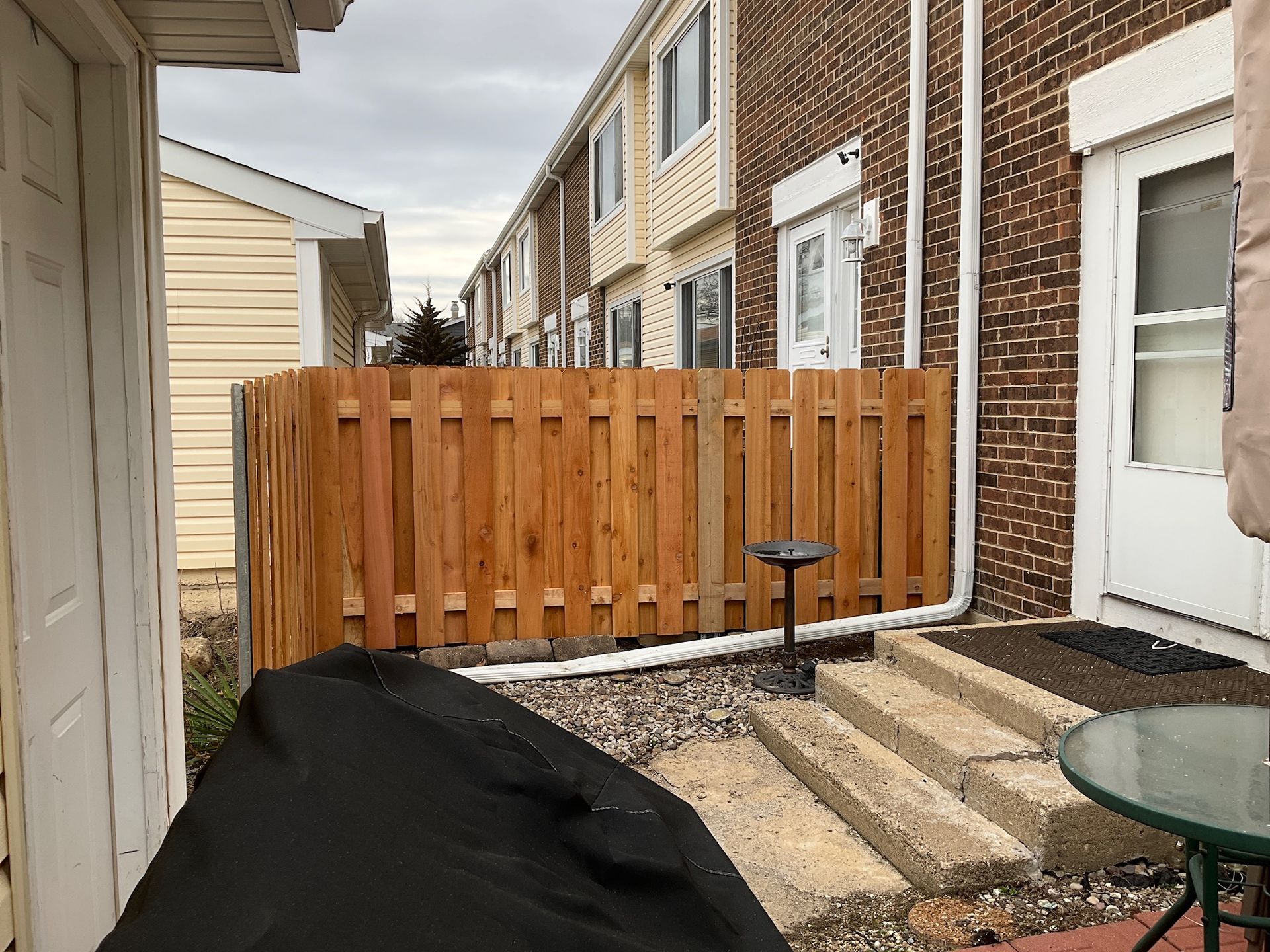 Wooden fence on a patio with stone steps, a bird bath, and several townhouses in the background.