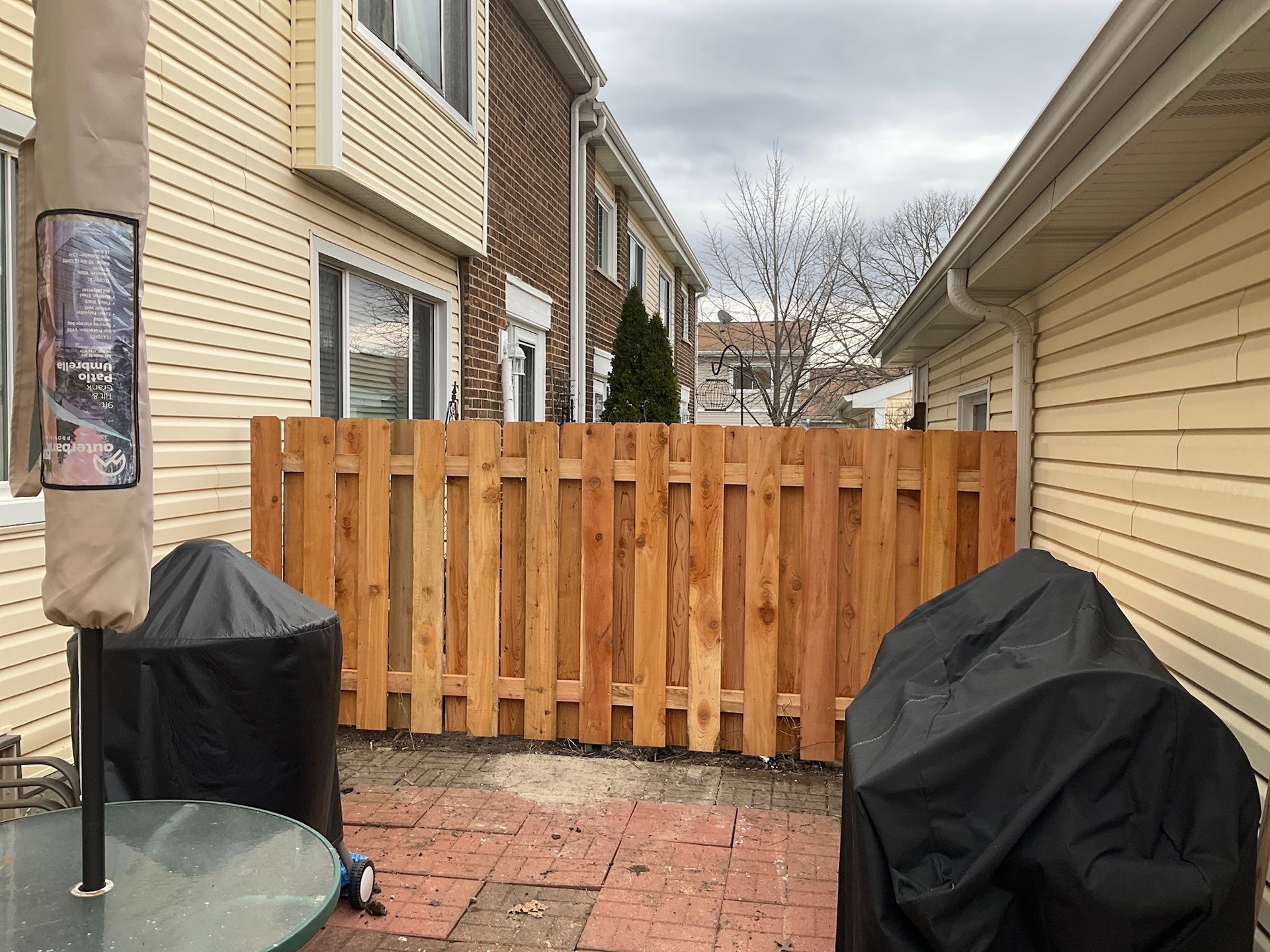 Wooden fence in a backyard with a red brick patio, flanked by buildings.