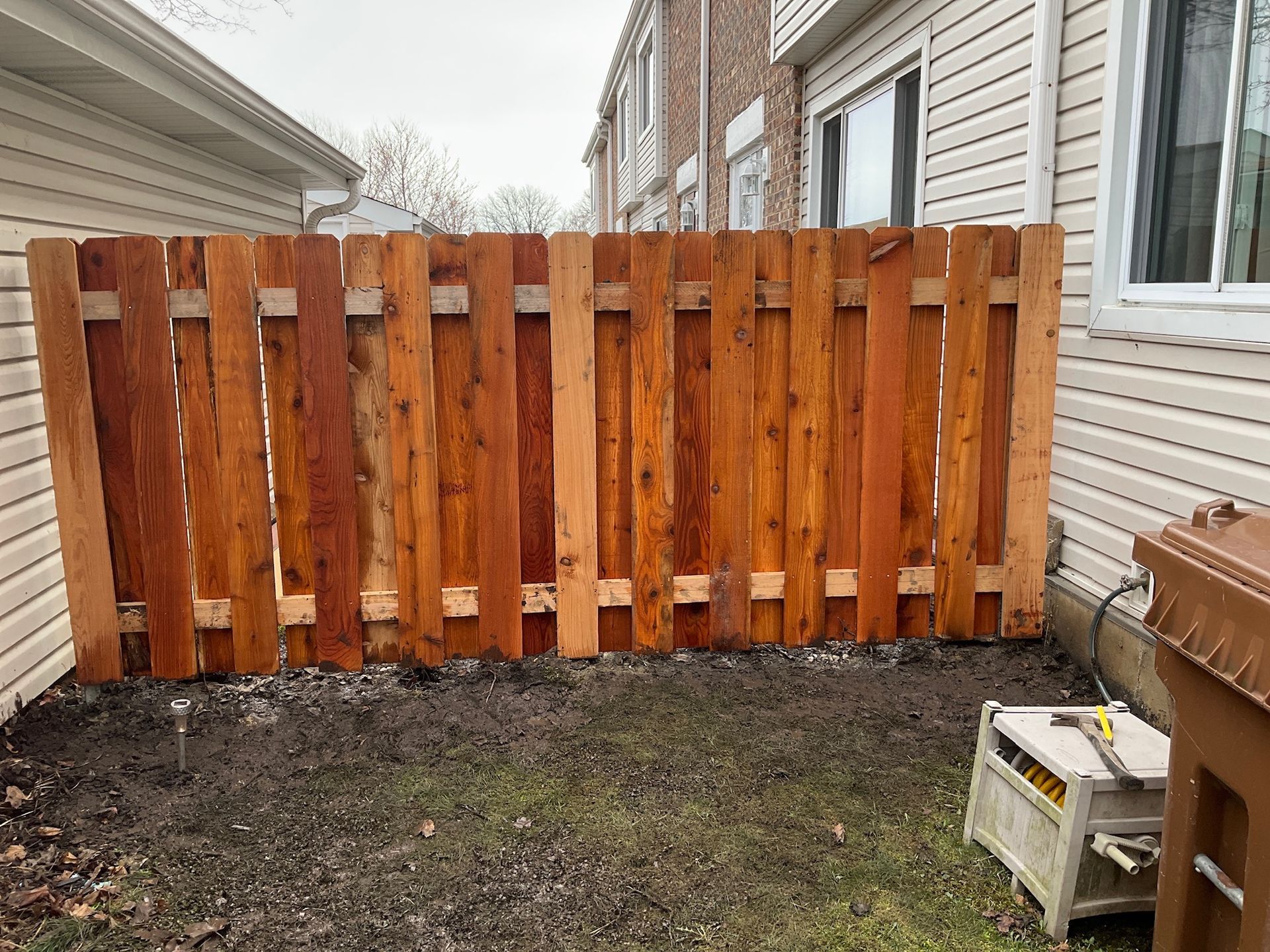 Wooden fence between two buildings; brown, vertical planks. Ground is muddy.