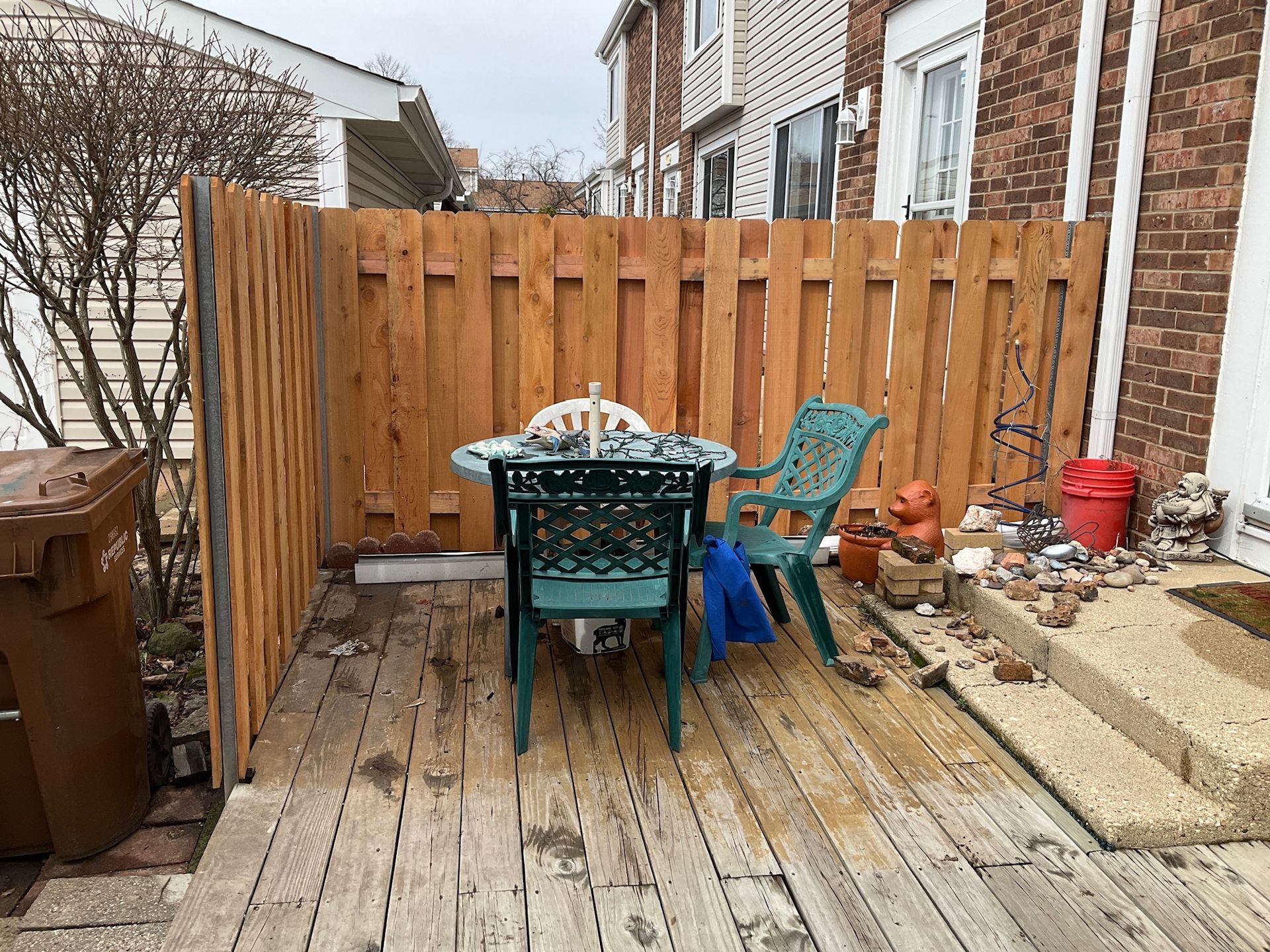 Wooden deck with small table and green chairs, enclosed by a wooden fence. Brown trash can on left.