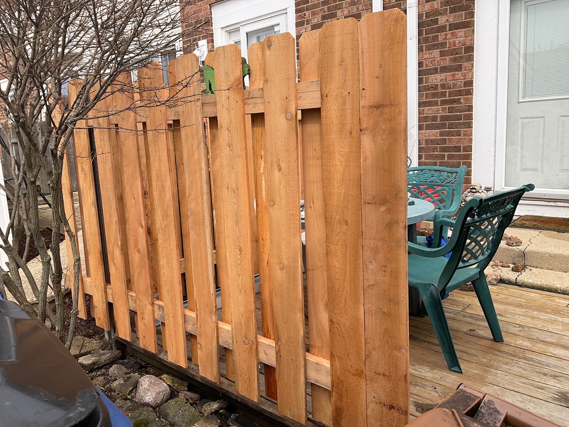 Wooden fence with vertical planks, brown stain, protecting a small patio with a green table and chairs.