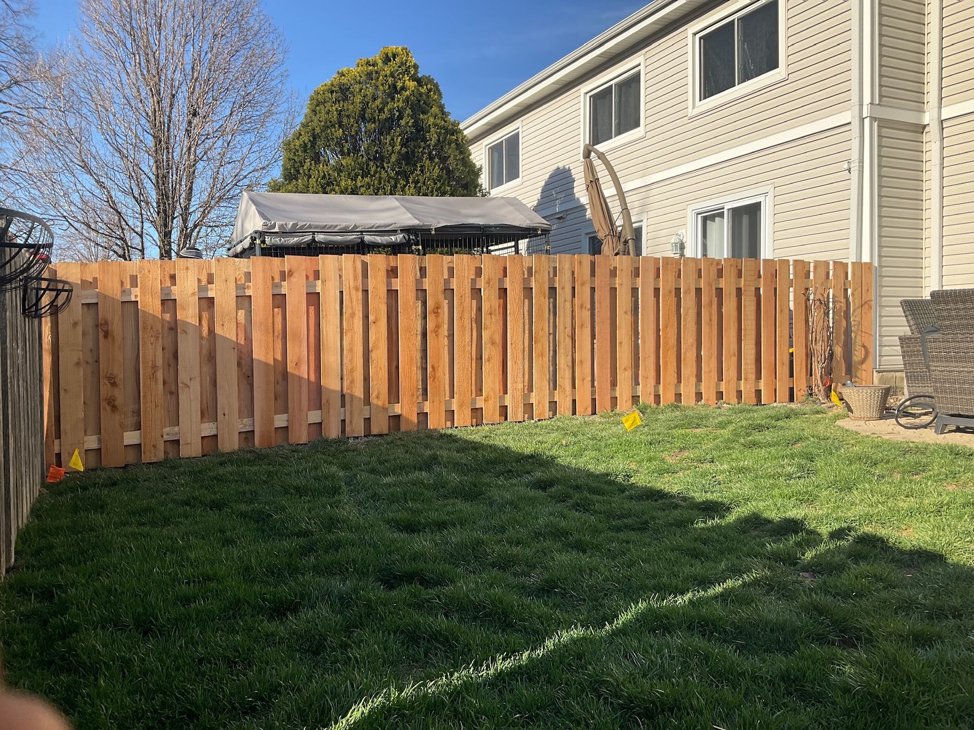 Wooden fence in a backyard with green grass, a townhouse in the background, and clear sky.