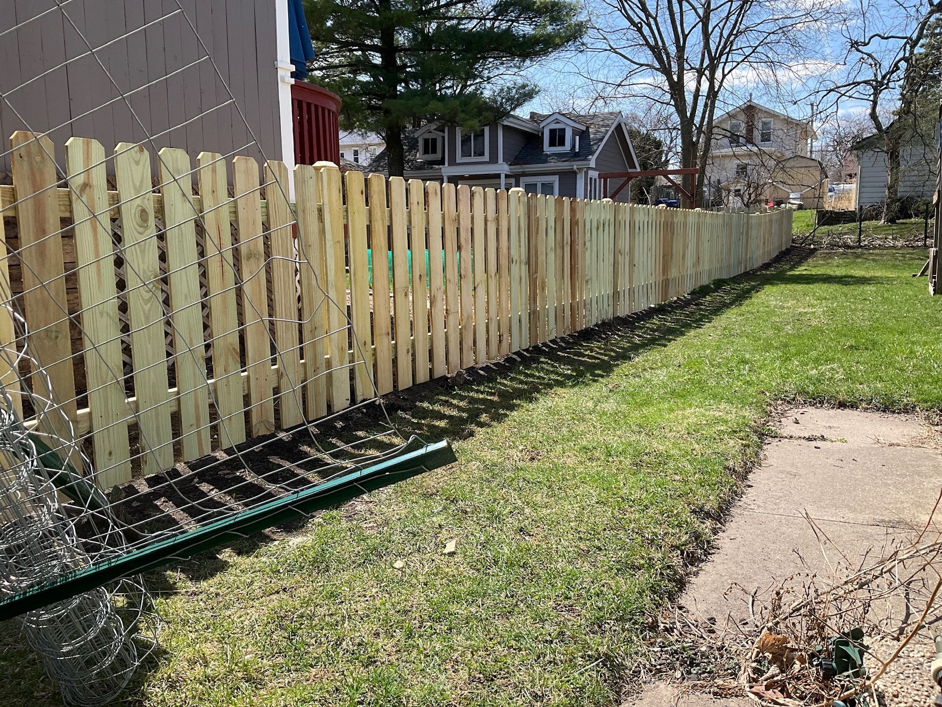 Wooden fence bordering a grassy yard on a sunny day. Houses are visible in the background.