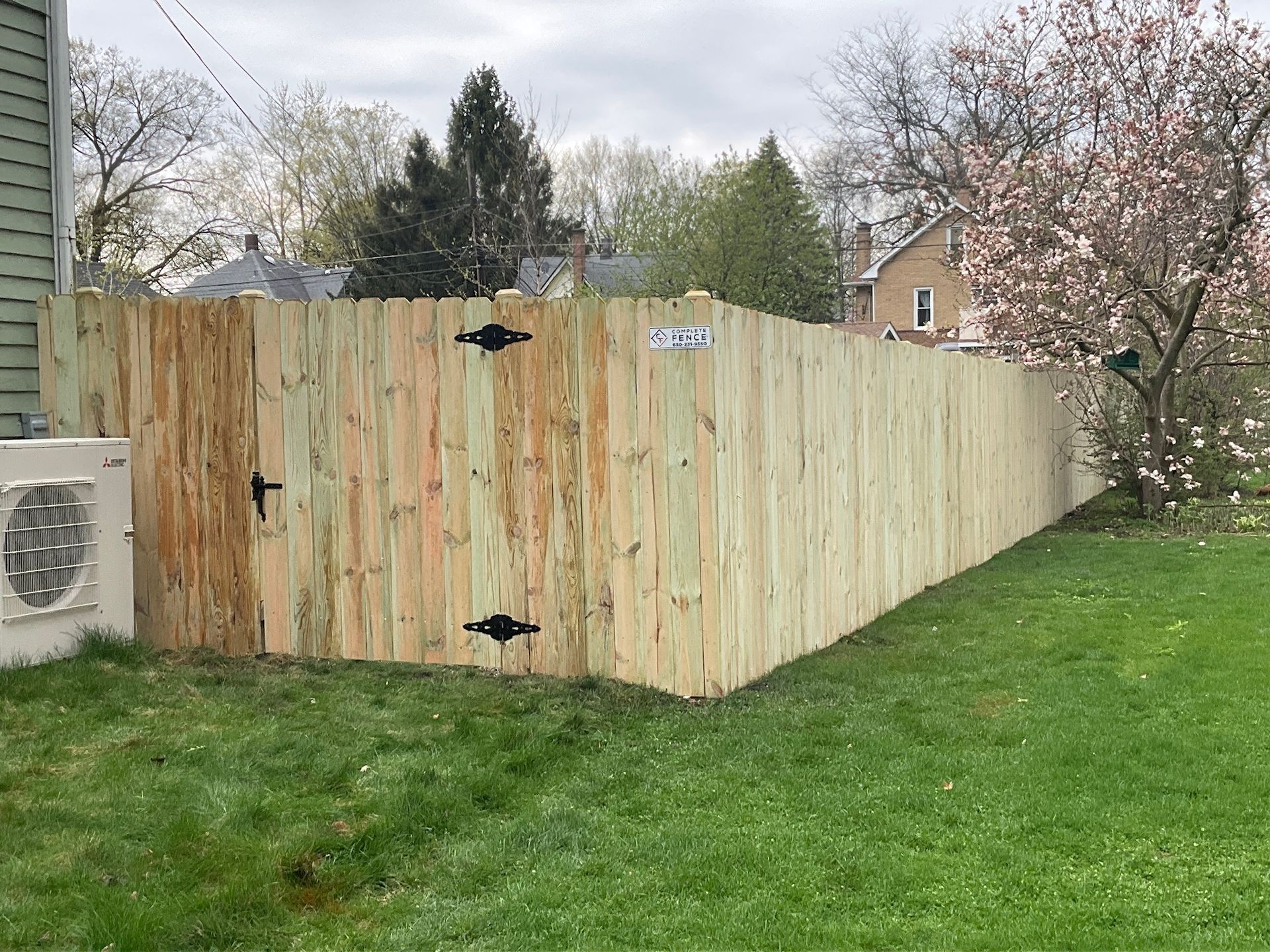 A newly built wooden fence encloses a green lawn, with a gate and a blooming tree in the background.