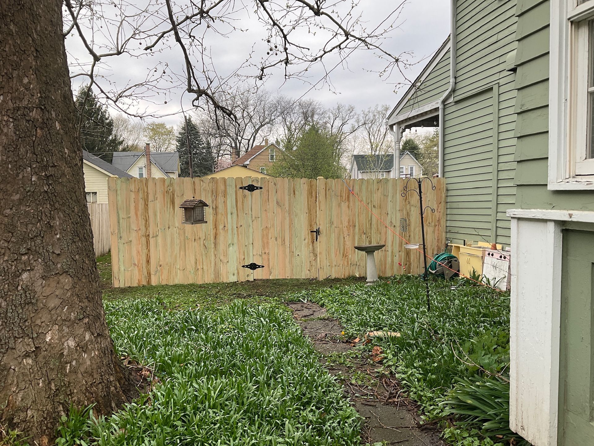 A wooden fence in a yard with a birdhouse and birdbath. Green grass and a tree are in the foreground.