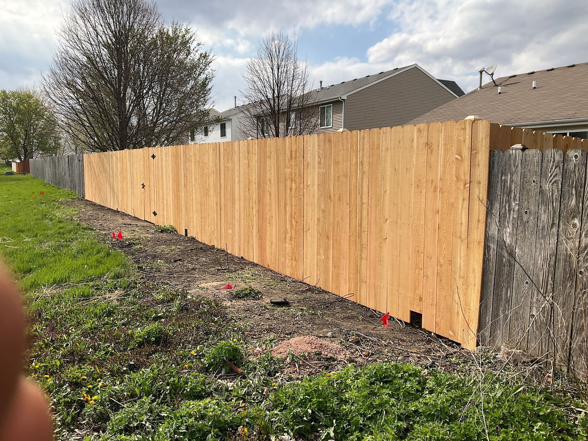 New wooden fence next to an old, weathered fence. Ground has grass and red flags. Cloudy sky.