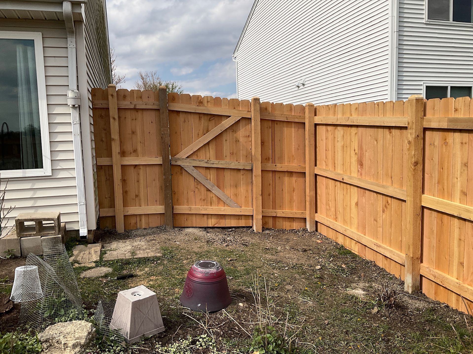 Wooden fence with a gate encloses a backyard; a white building is on the left, and another on the right.