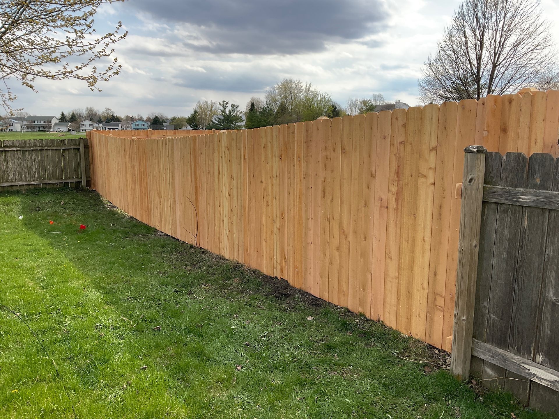Wooden fence in a grassy yard, partially constructed next to an older fence, under a cloudy sky.