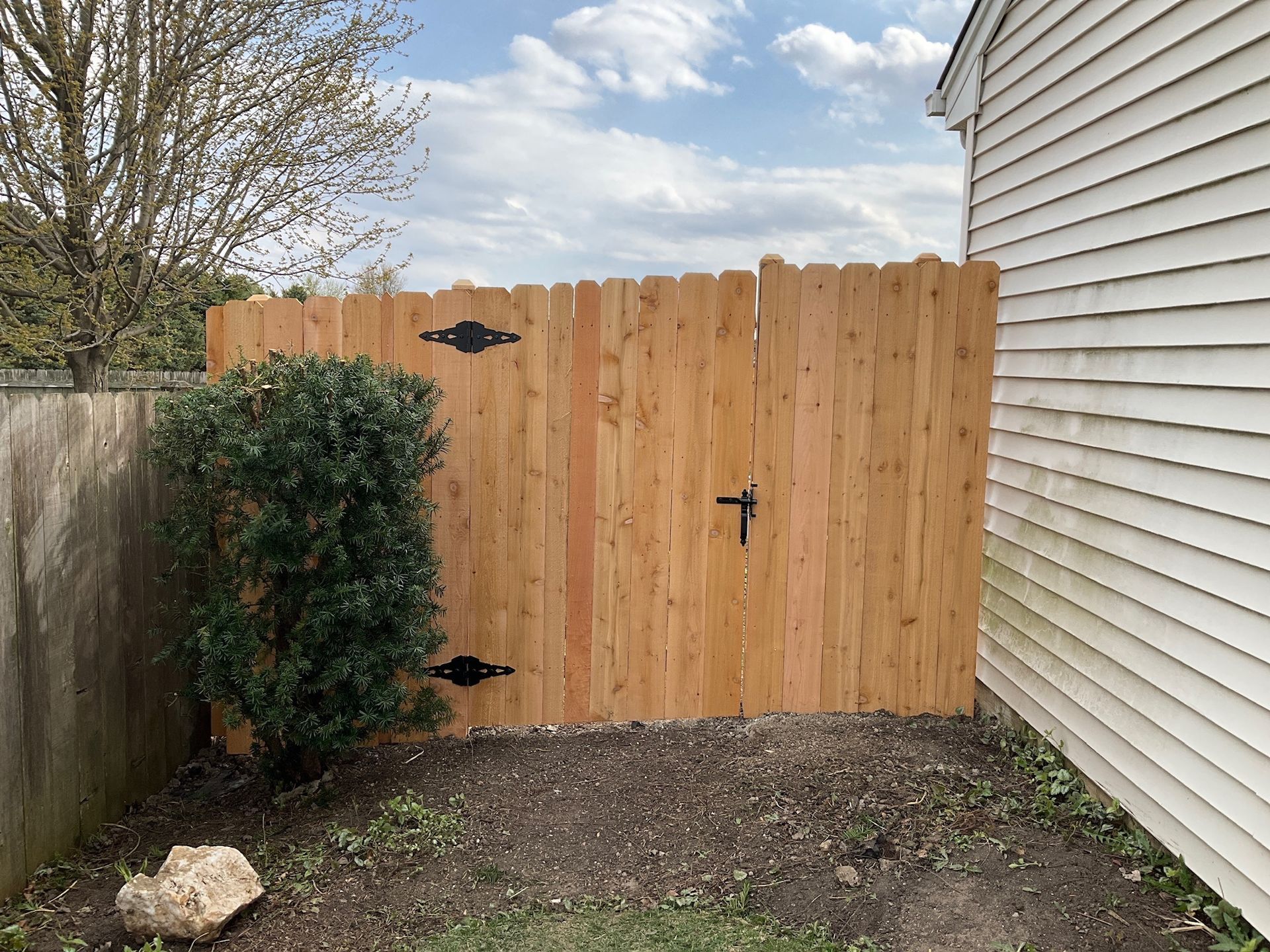 Wooden gate with black hardware between a concrete wall and a white-sided house, with sky in background.