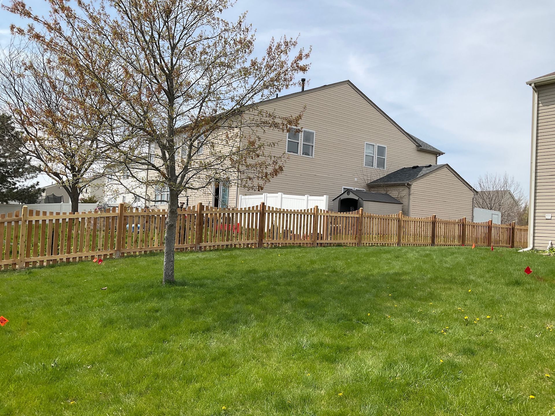 Wooden picket fence encloses a green lawn with a tree and a two-story beige house under a cloudy sky.