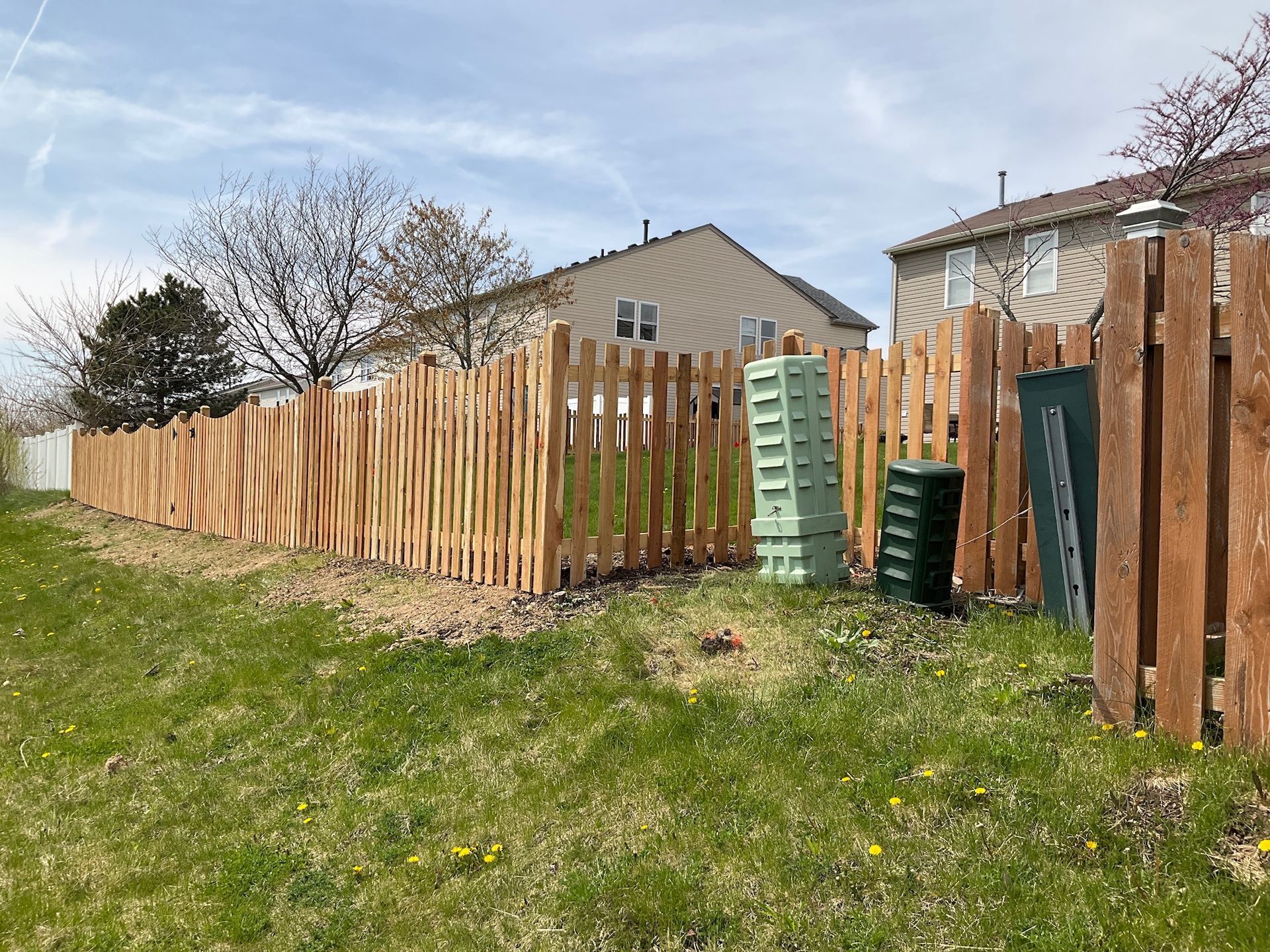 Wooden fence enclosing a grassy yard; houses in the background; green compost bins.