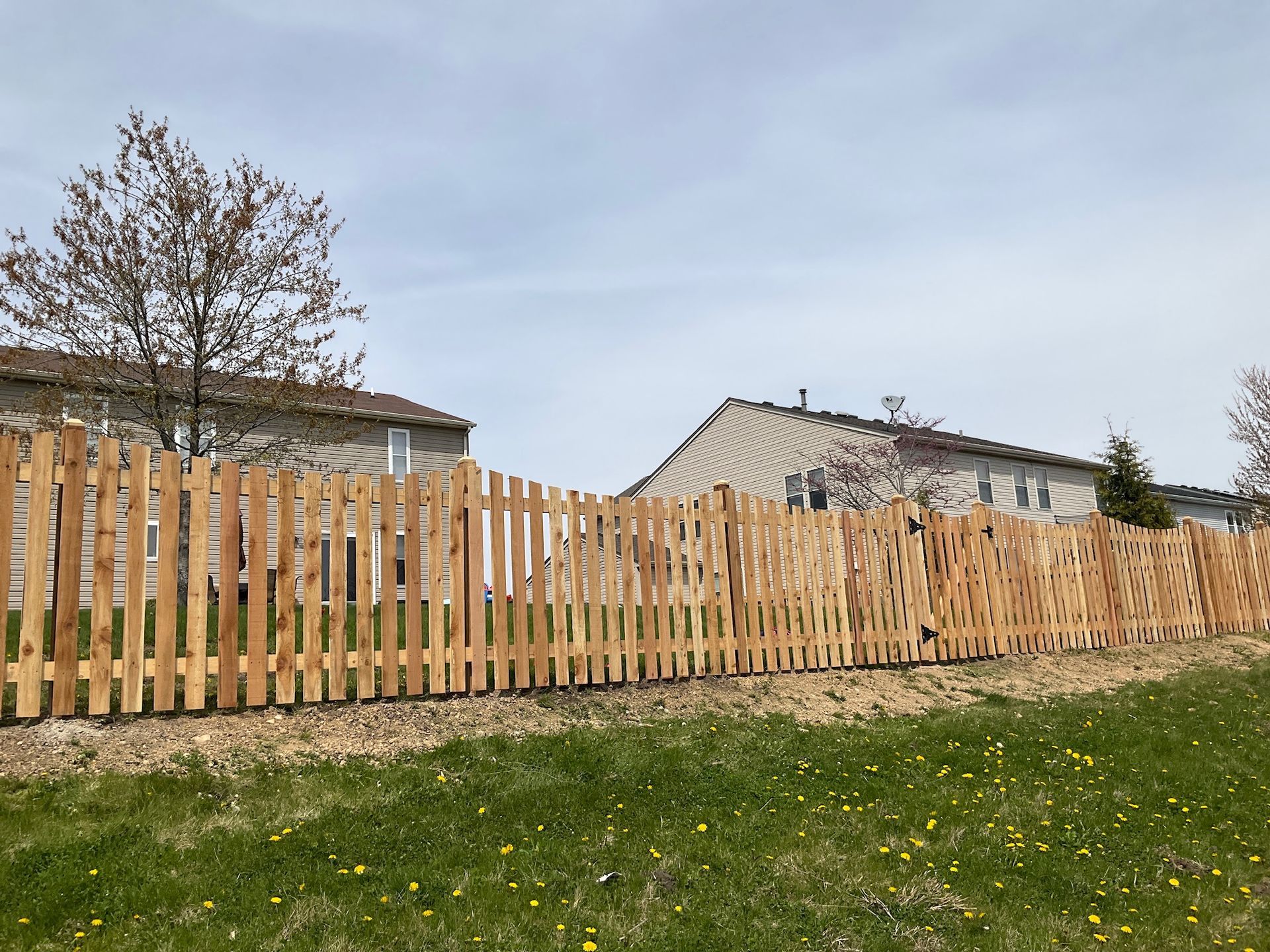 Wooden fence along green grass with two buildings in the background and a cloudy sky.