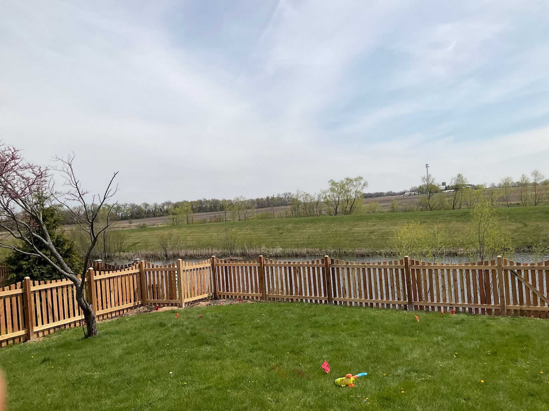 Lawn and wooden fence in a yard with a view of a field and cloudy sky.