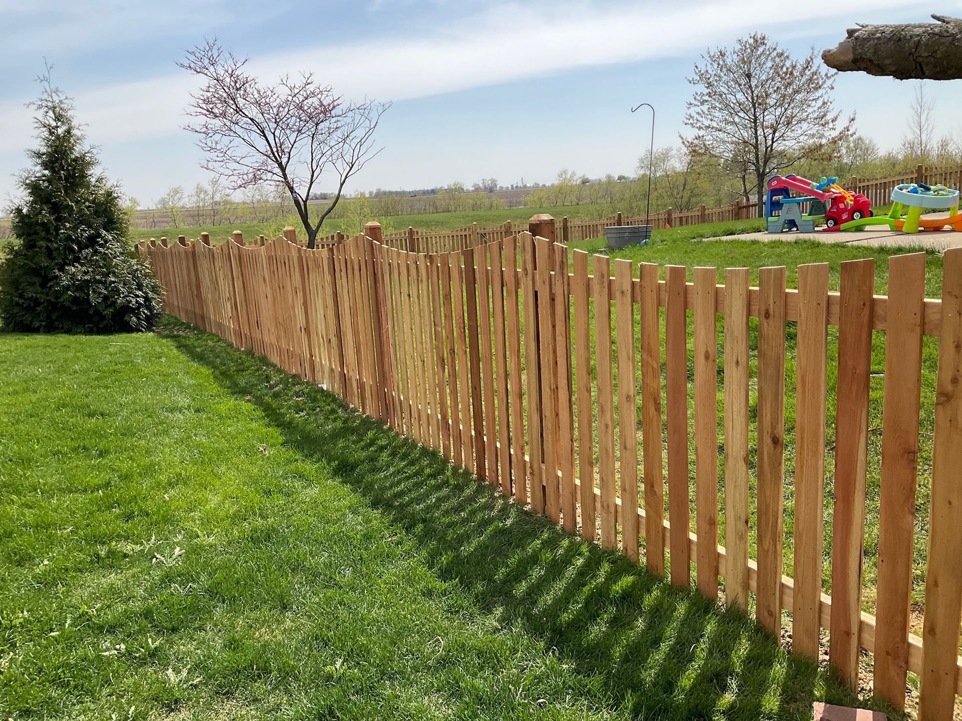 Wooden picket fence on green lawn, under blue sky; playground toys in background.