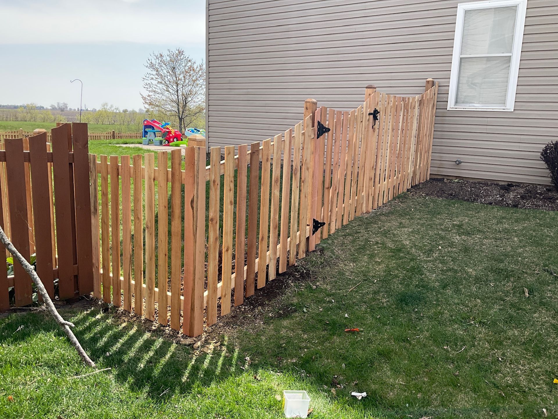 Wooden picket fence in a yard, beside a house. Brown stained posts cast shadows on the grass.