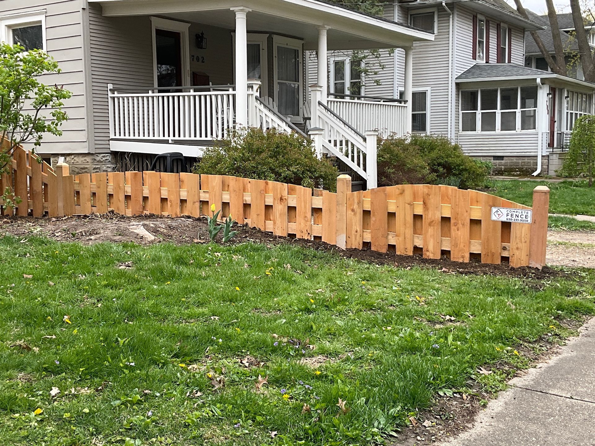 Wooden picket fence in front of a light-colored house with a porch and steps. Green grass and sidewalk.