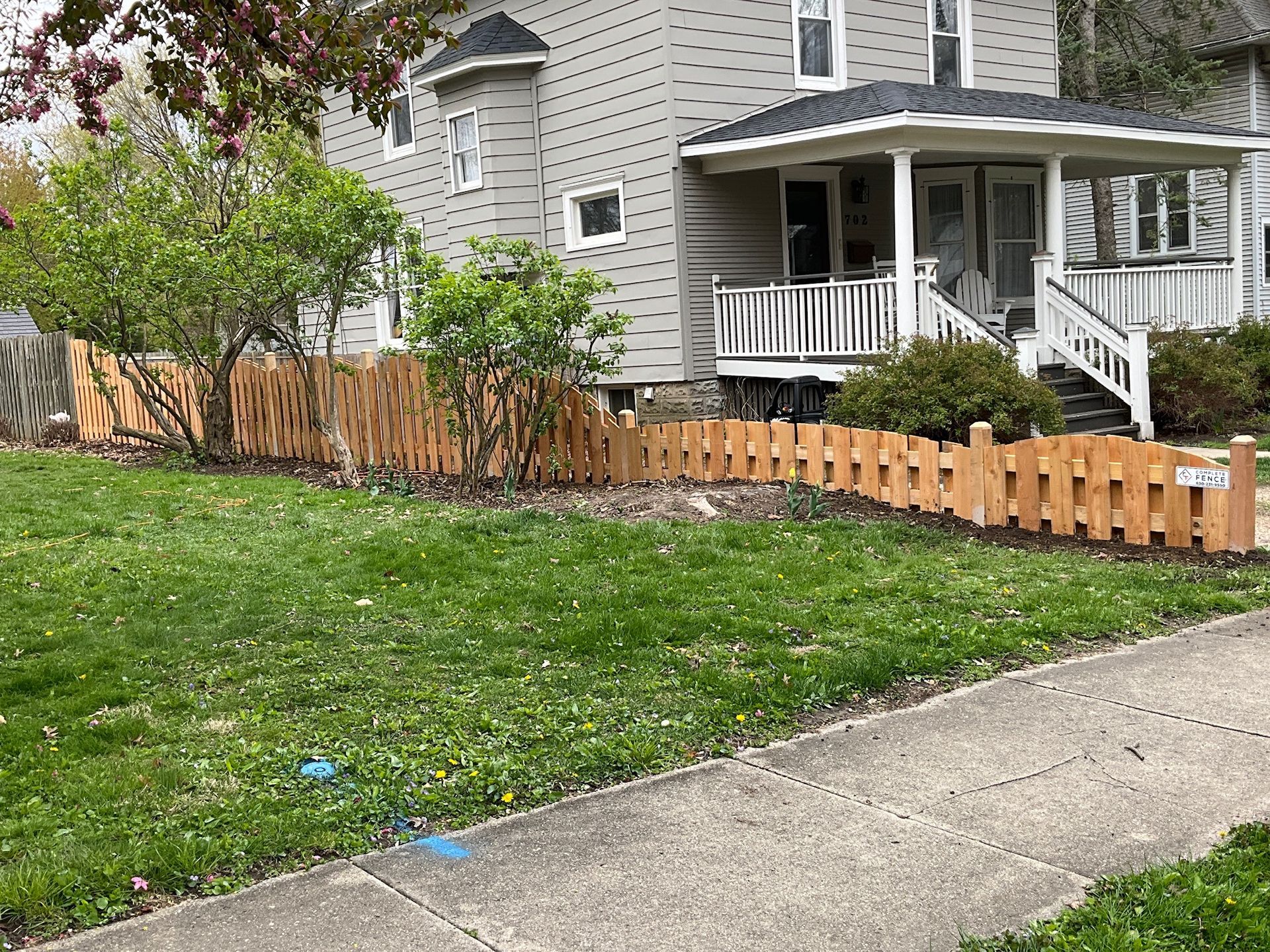 A light-colored wooden picket fence in front of a house, with a green lawn and sidewalk.