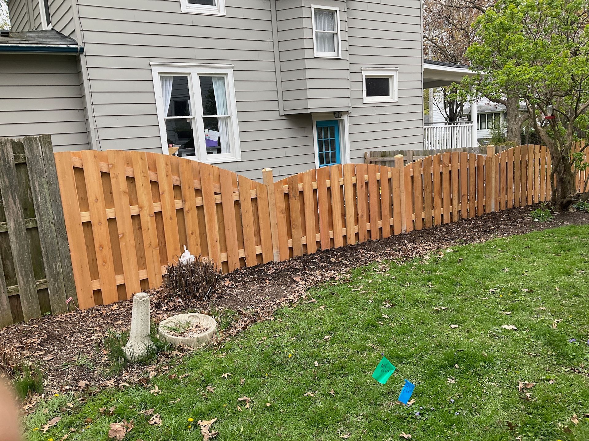 Wooden fence alongside a grey house with windows. Green grass and yard.