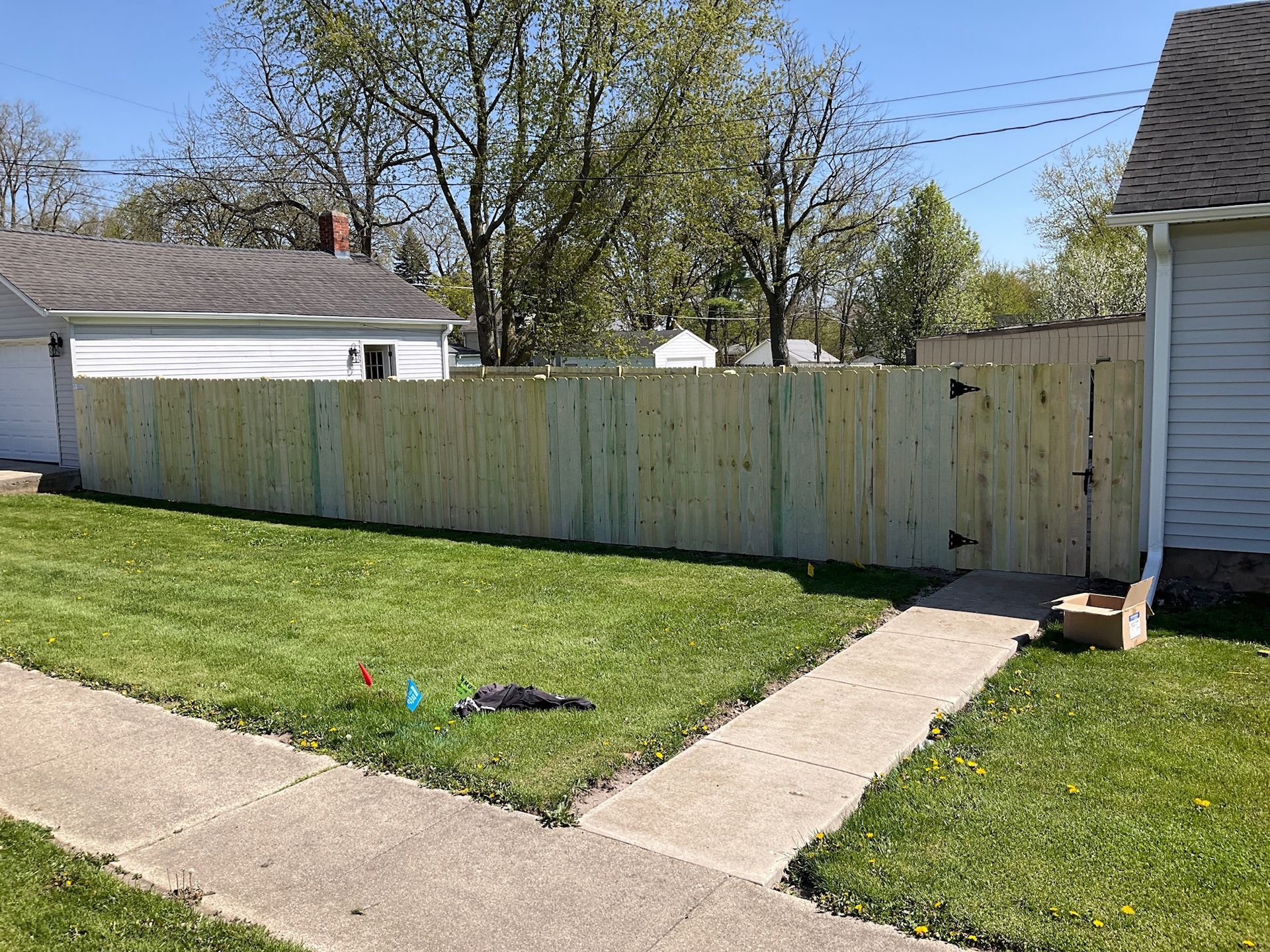 Wooden fence bordering a grassy yard, sidewalk in the foreground, houses and trees in the background.