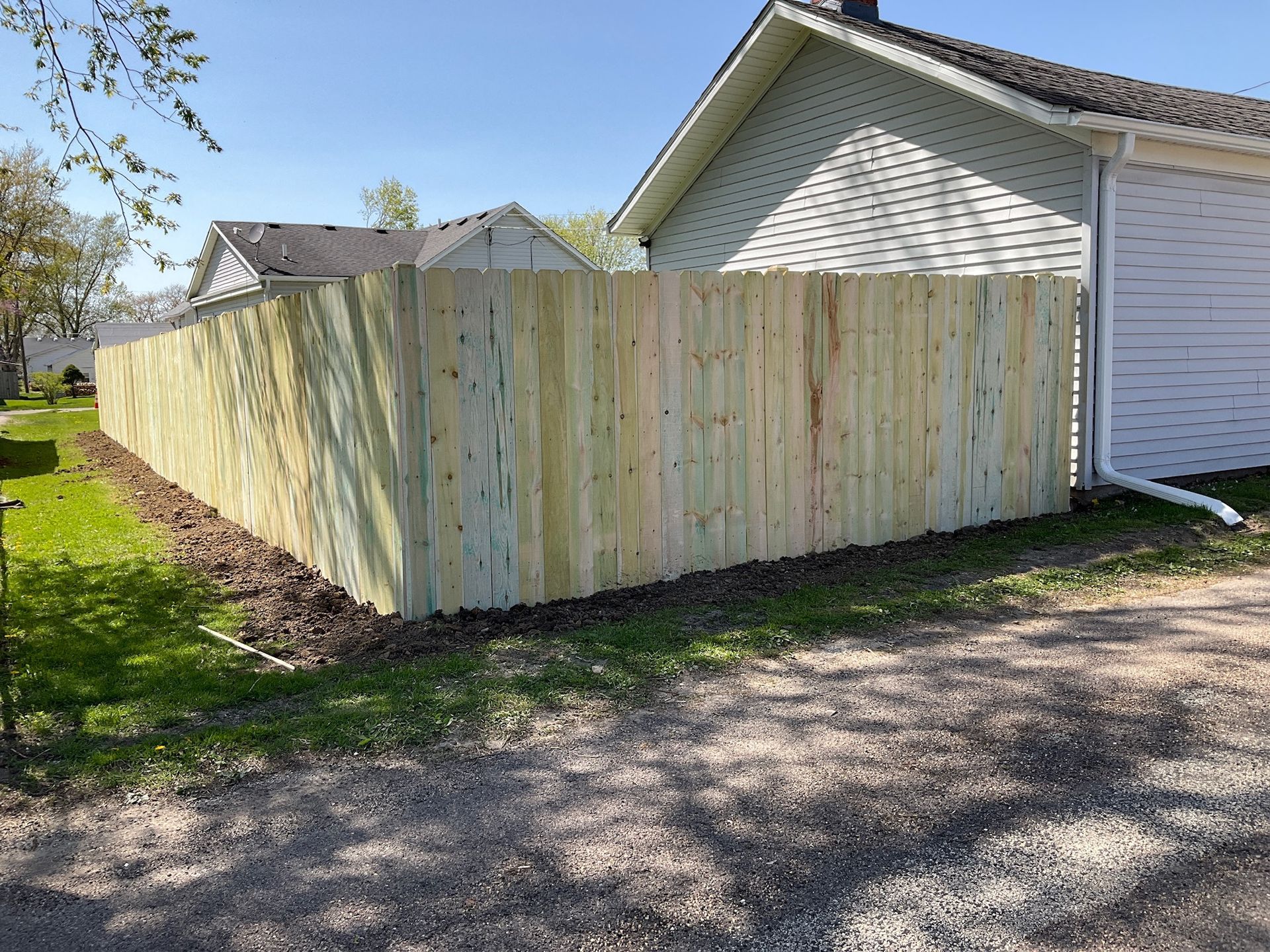 Wooden fence next to a house with a gravel driveway and grass, on a sunny day.